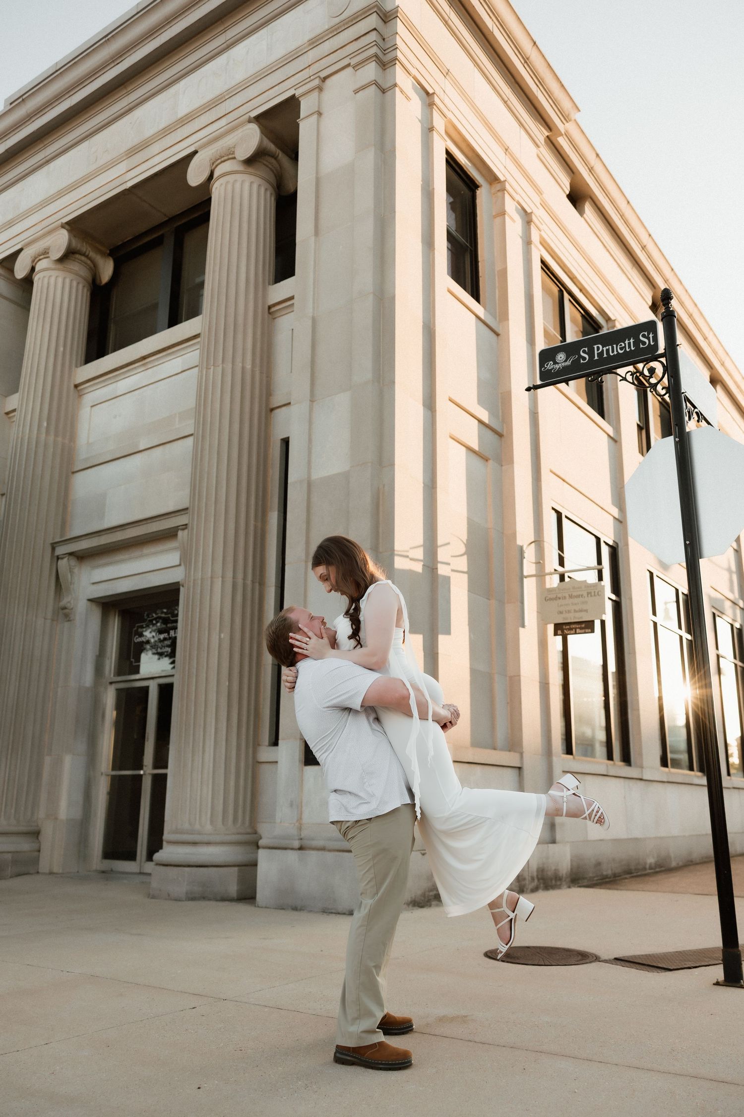 A man lifts his partner in a white dress outside a classic columned building at golden hour.