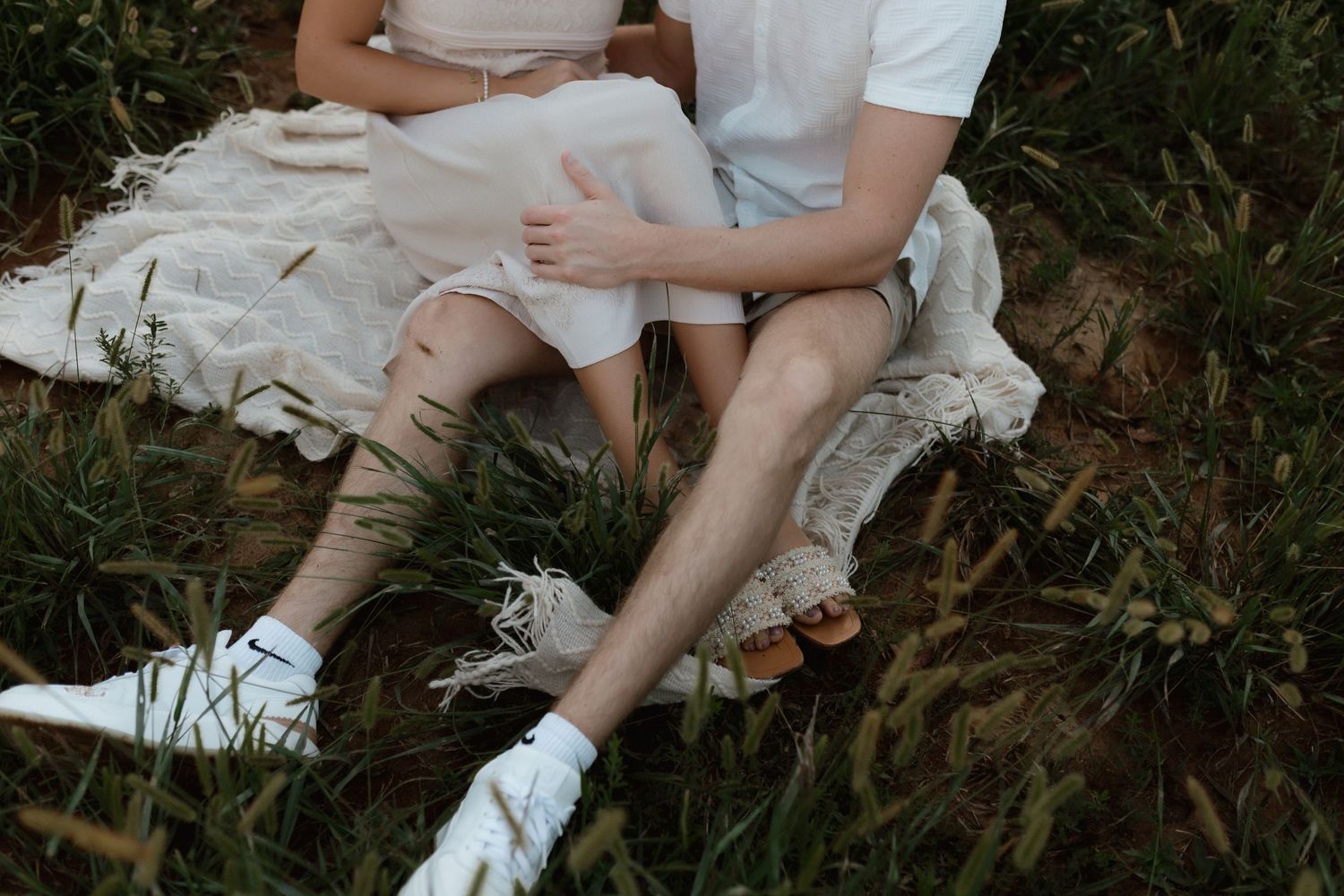 Close-up of couple sitting in tall grass, wearing white outfits during outdoor engagement session.