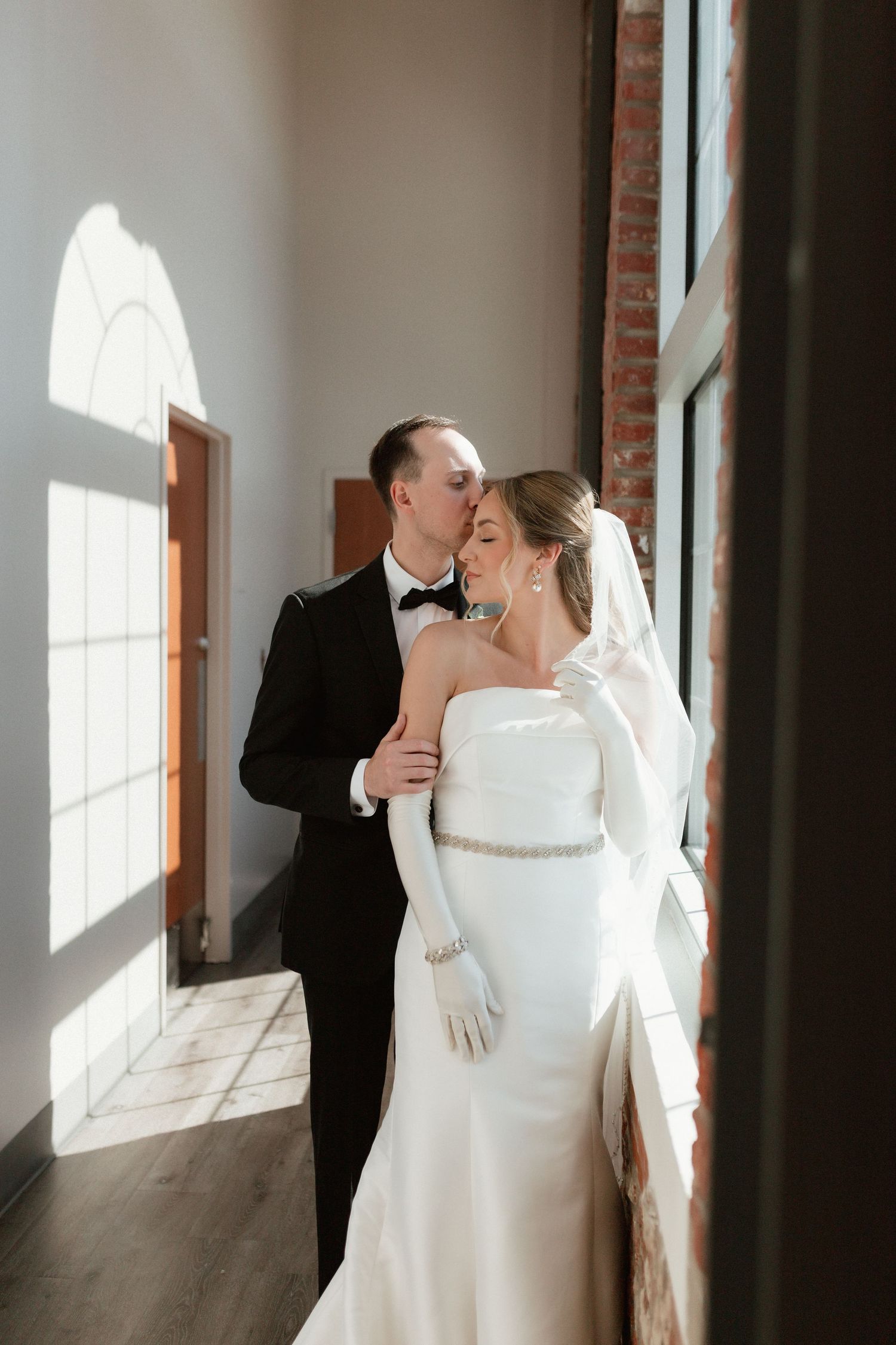 Groom tenderly kisses bride on the cheek near sunlit windows in brick venue.