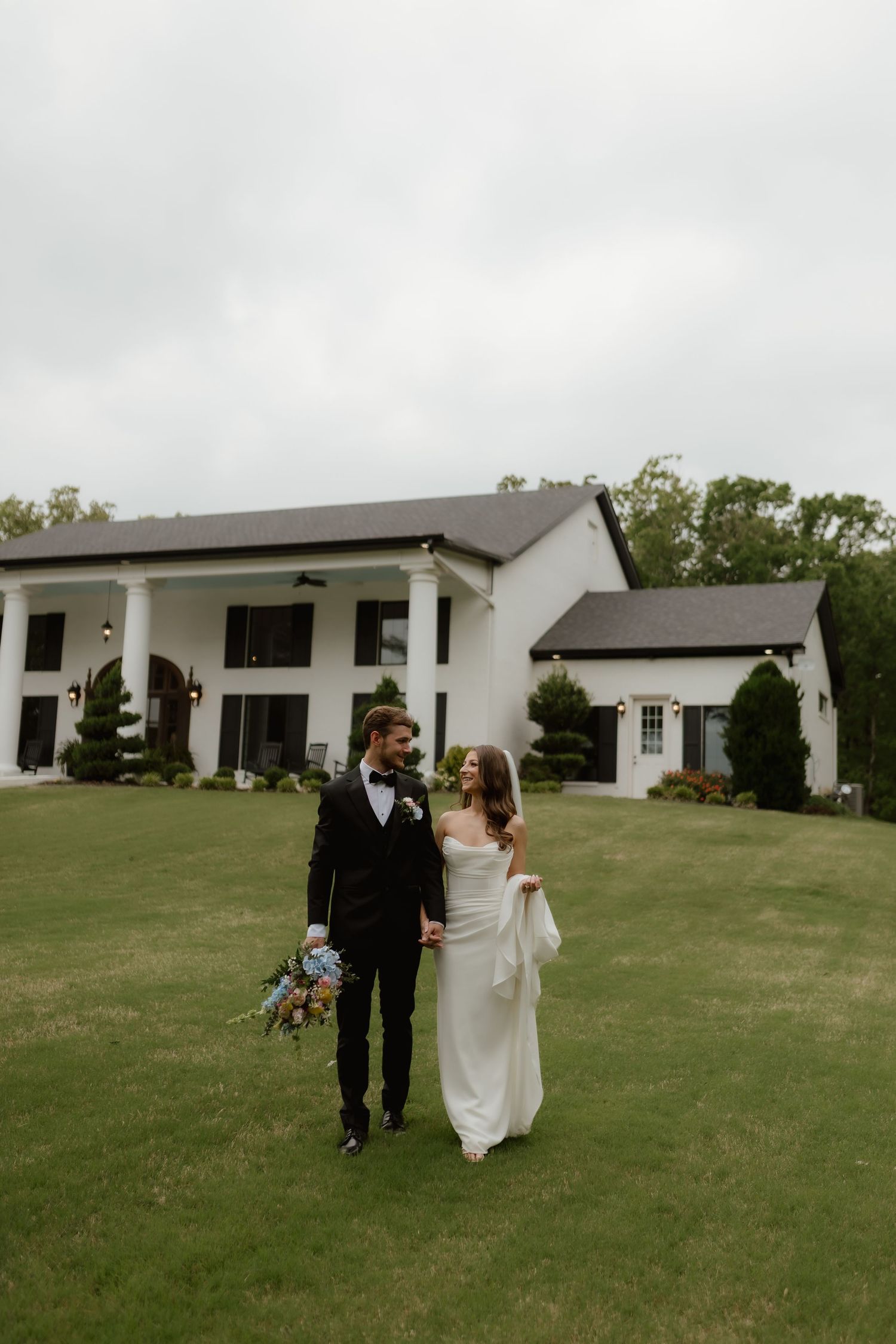 Bride and groom walk hand in hand across green lawn in front of elegant white estate.