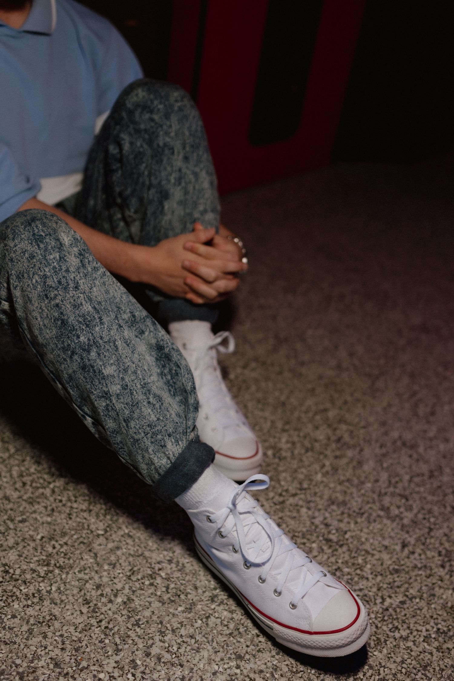 Person sitting on floor in acid wash jeans and white high-top Converse sneakers.