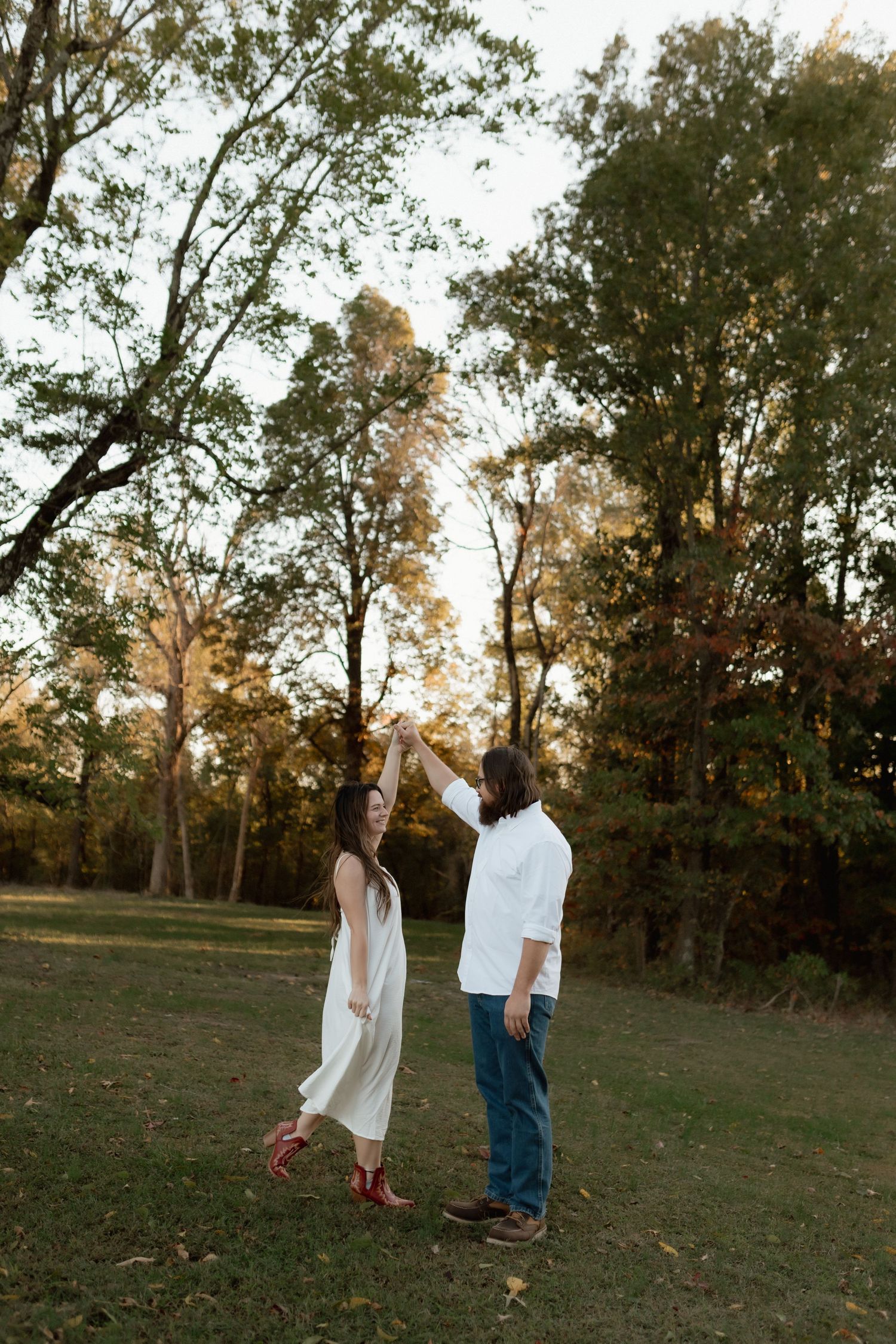 A couple dances together in a park at sunset, man spinning woman in white dress and red cowboy boots on green grass.