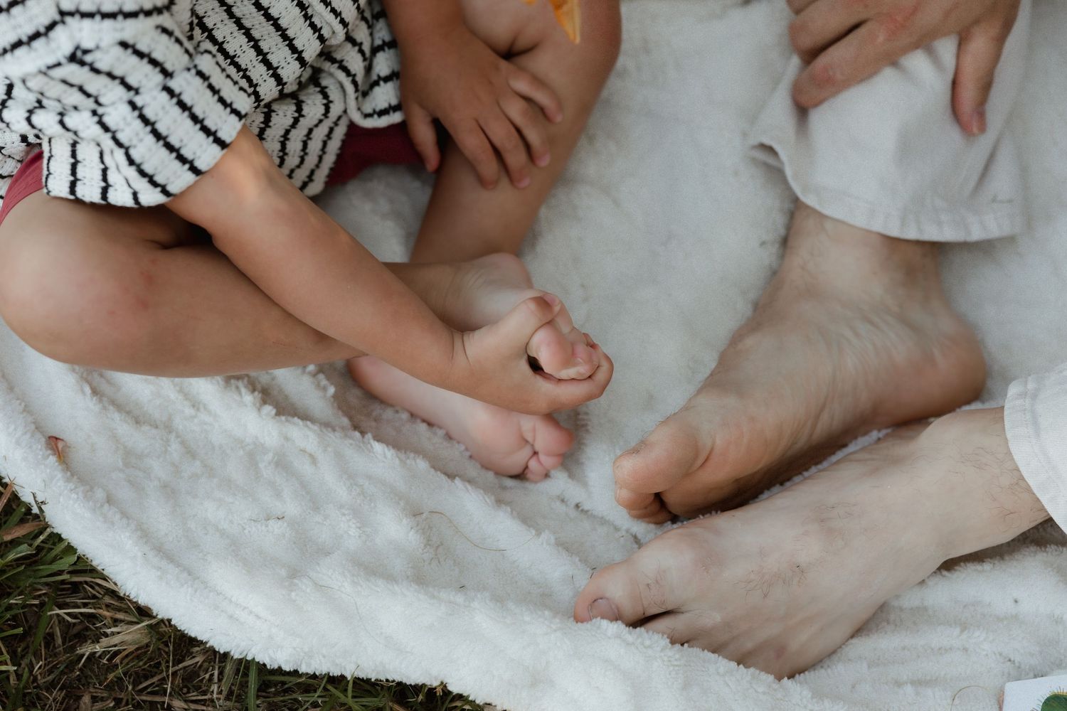 Close-up of adult and toddler bare feet resting together on soft white blanket outdoors.