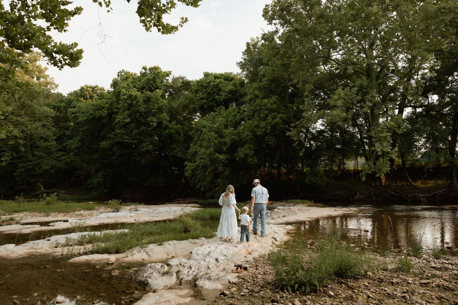 Family of three standing on rocky riverbank surrounded by lush green trees in natural setting.