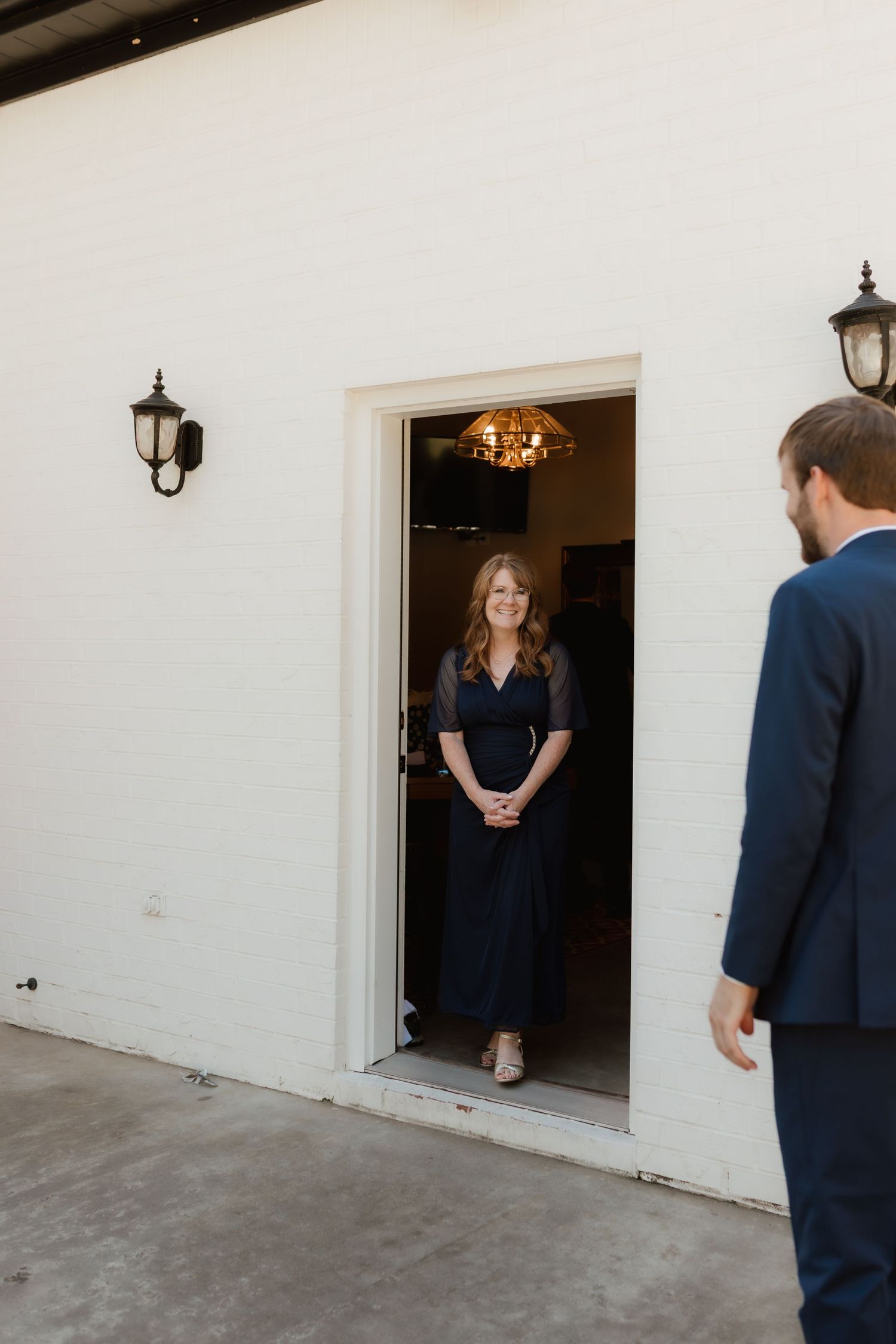 Woman in navy dress standing in a doorway smiling at a man in a suit outside a white building.