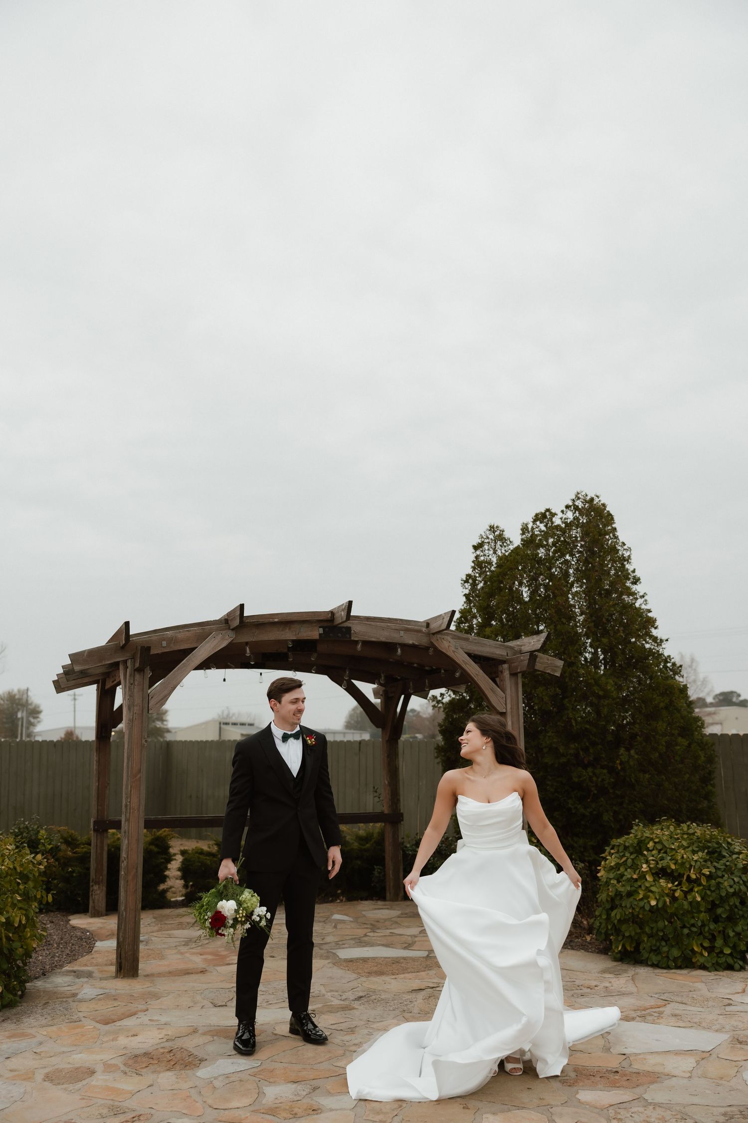 Bride lifting white gown while groom in black tuxedo walks beside wooden pergola on overcast day.