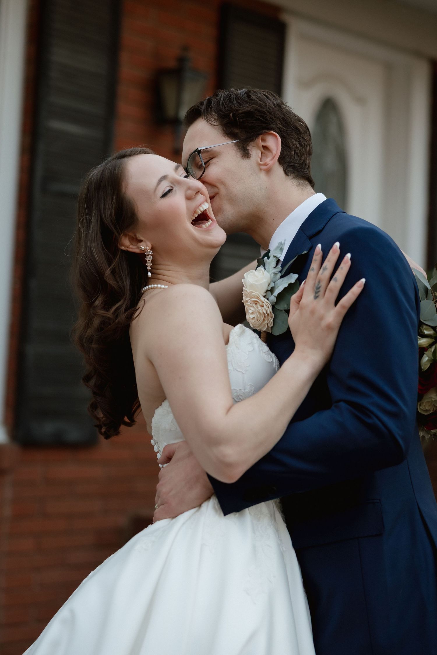Laughing bride in strapless gown embraced by groom in navy suit outside brick building.
