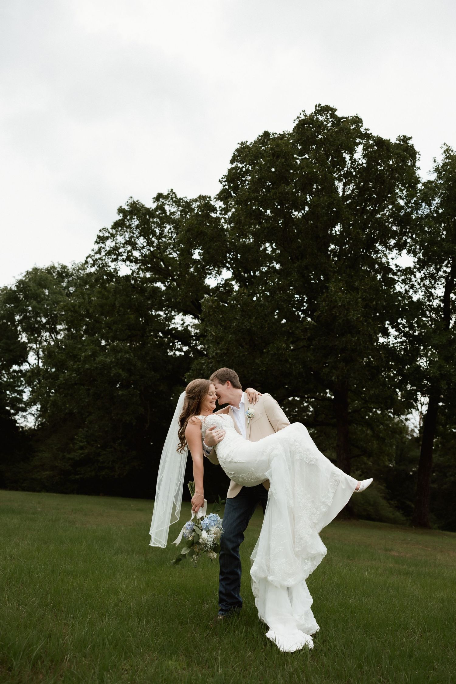 Groom lifts and kisses bride in white lace gown and veil in an outdoor field.