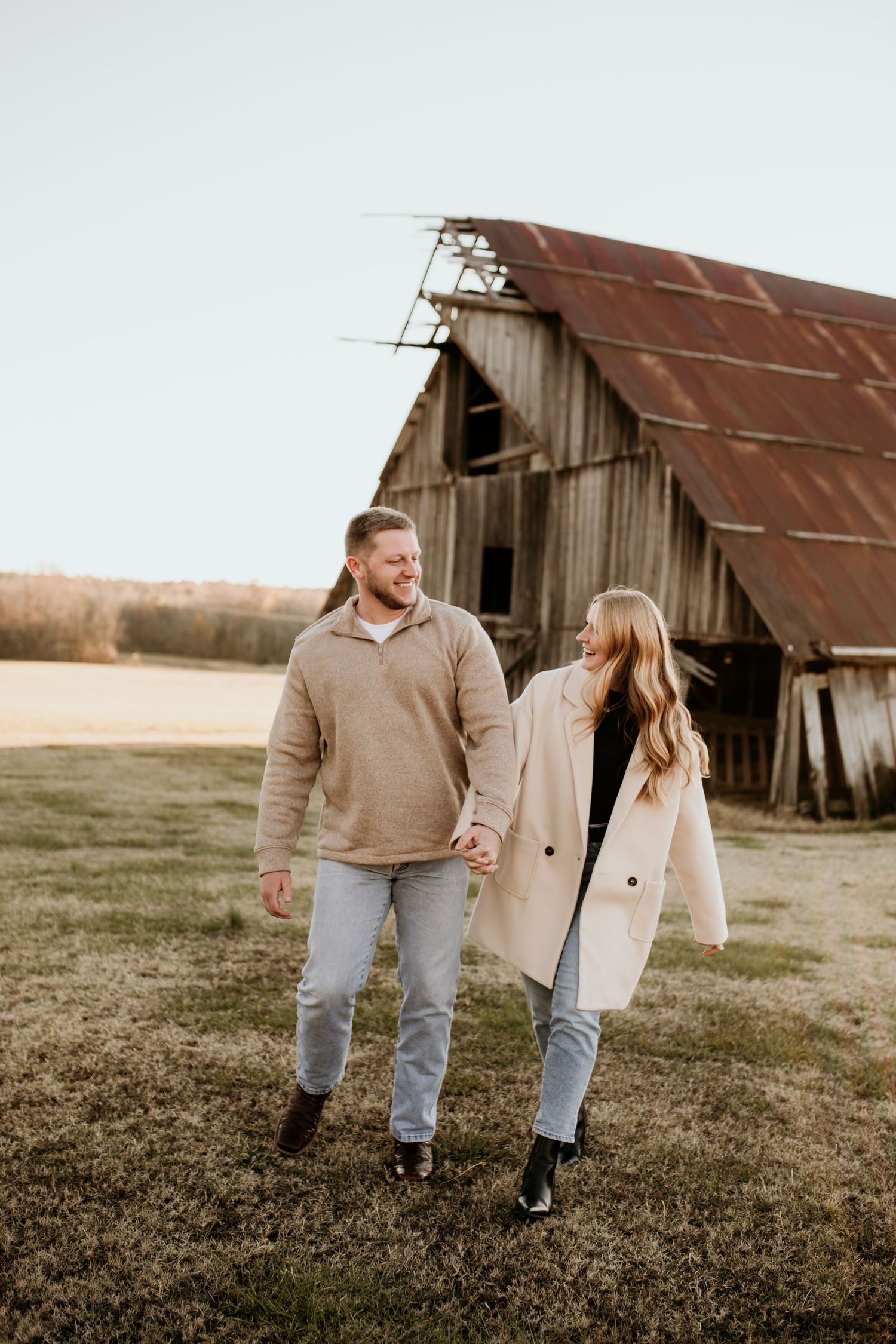 Happy couple holding hands walking together near weathered barn with rusty metal roof in open field.
