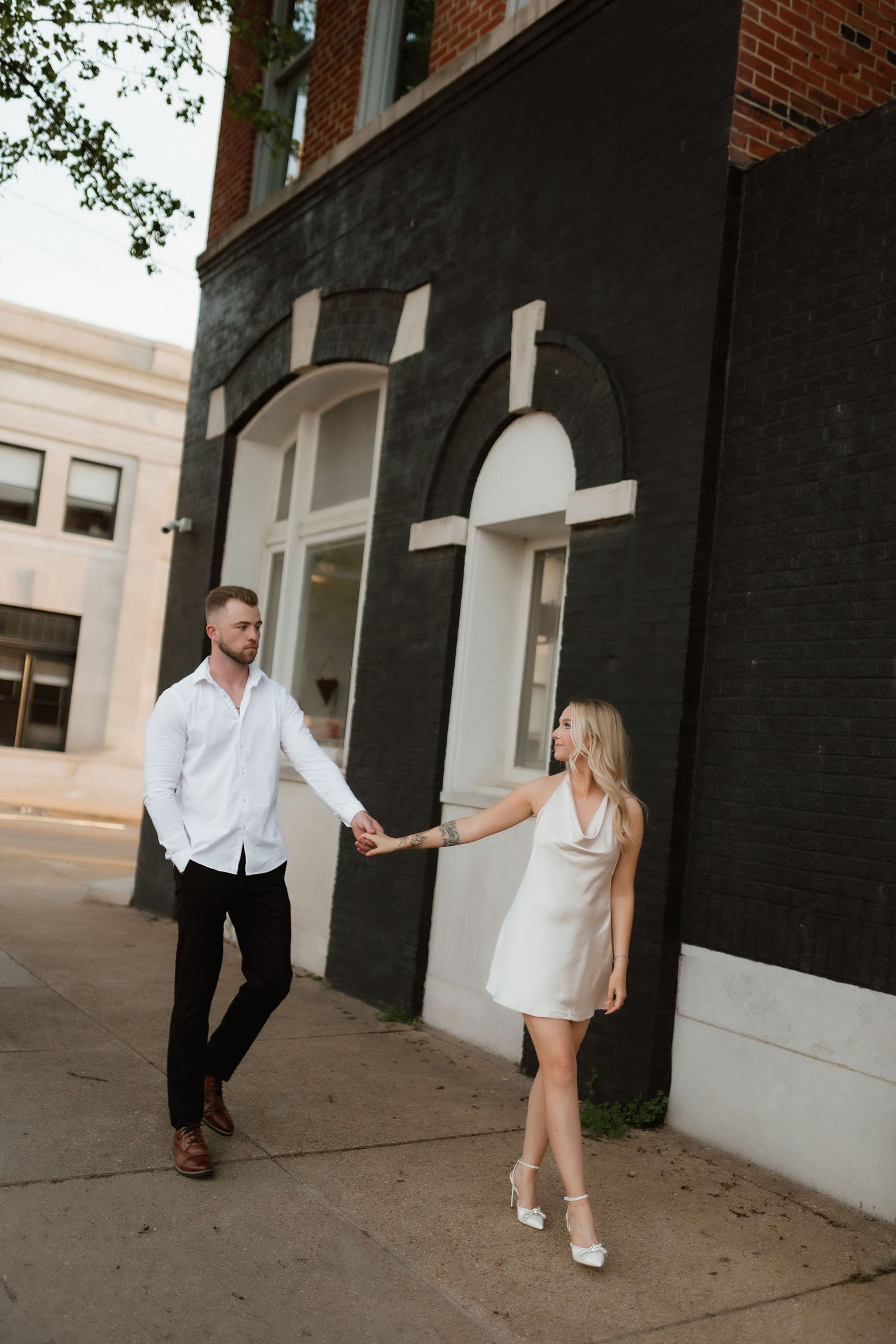 An elegant couple holds hands walking past a black building with arched windows at sunset.