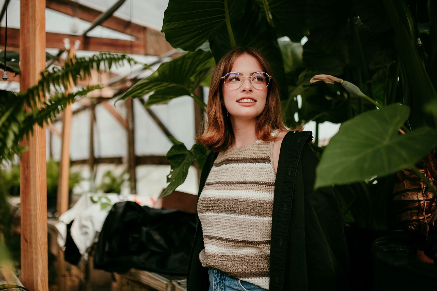 Young woman with glasses and striped top explores a lush tropical greenhouse filled with large green plants.