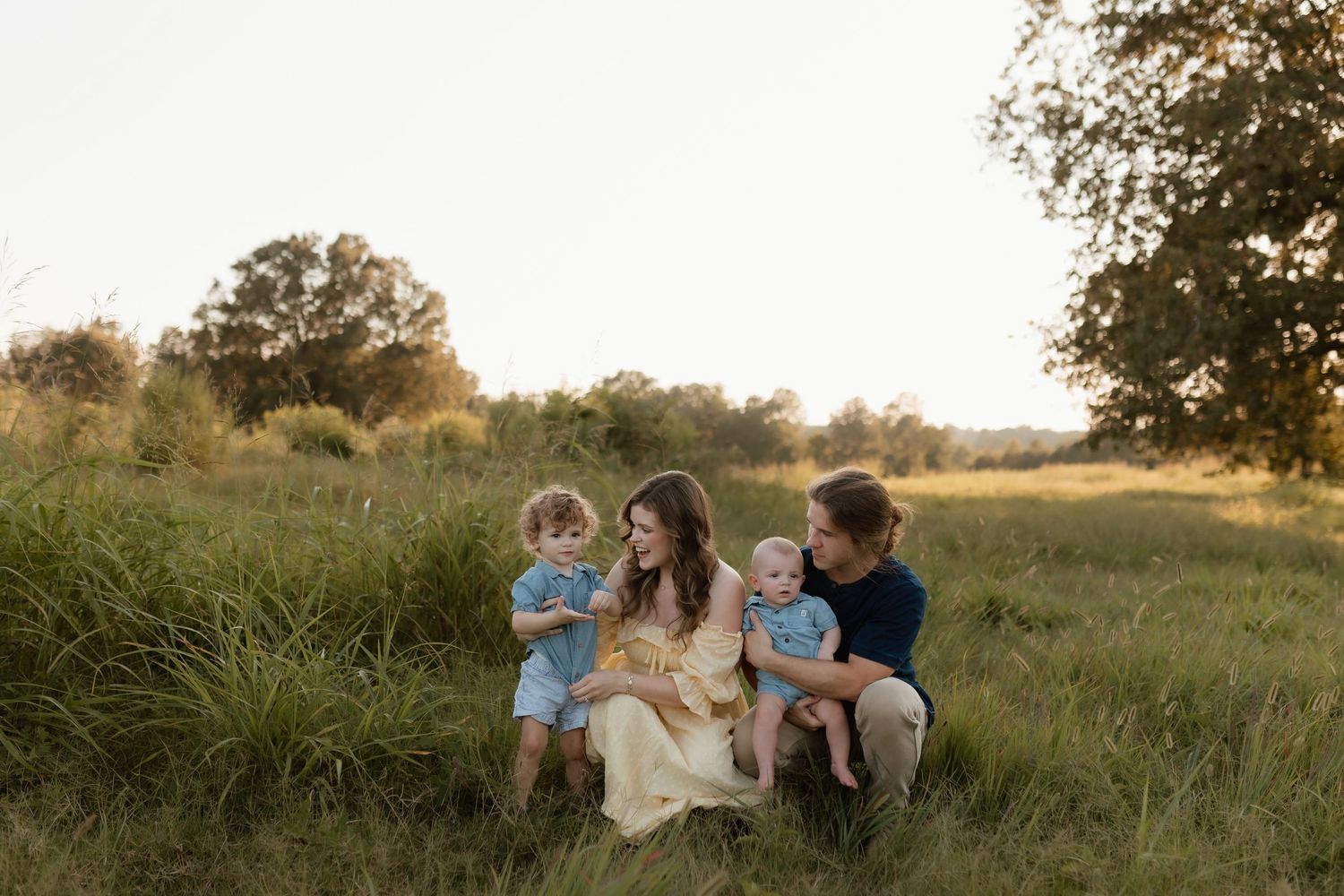 Family of four sitting in golden field at sunset, mother in yellow dress with toddler and baby.