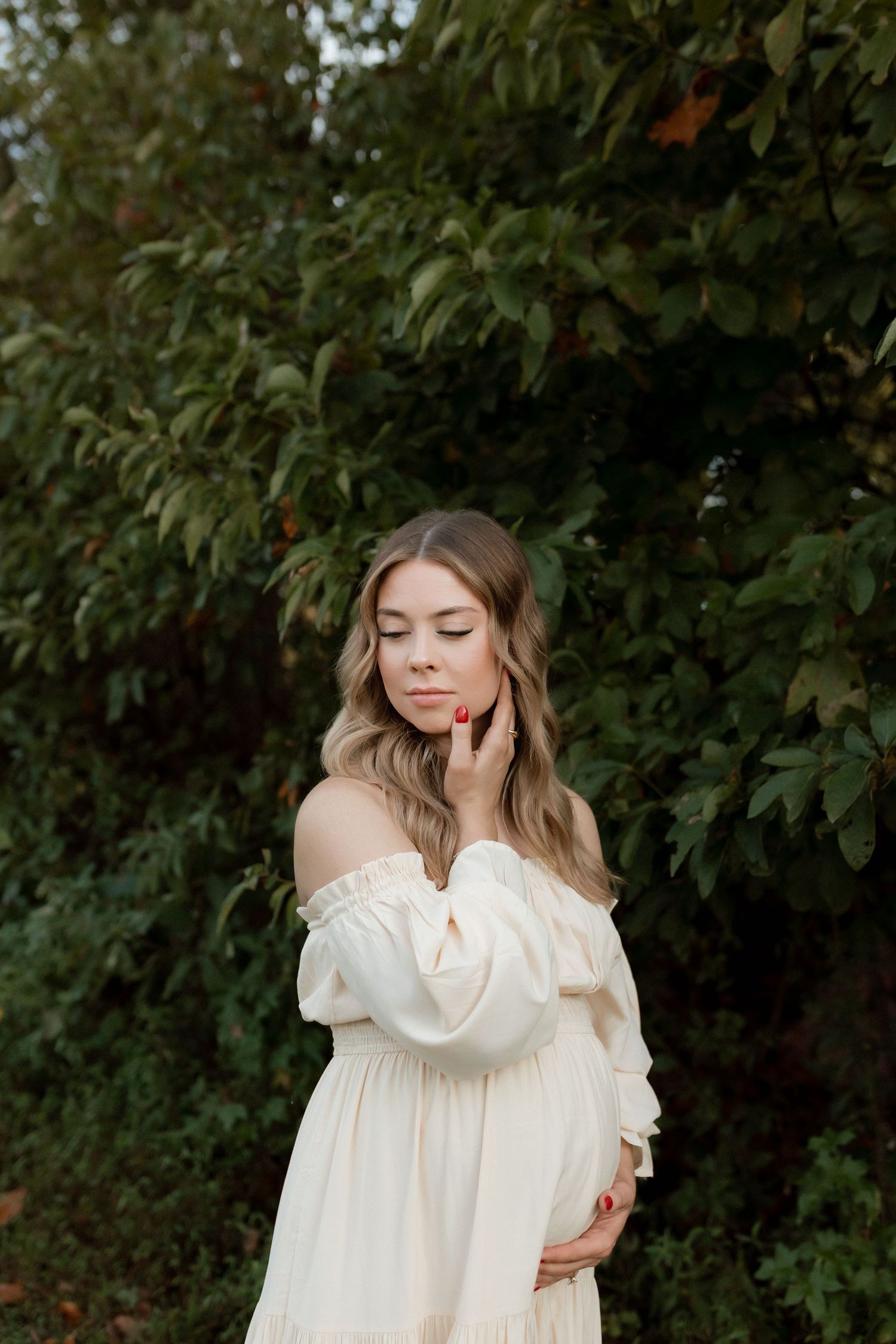 Serene pregnant woman in cream off-shoulder dress posing with eyes closed near green foliage.