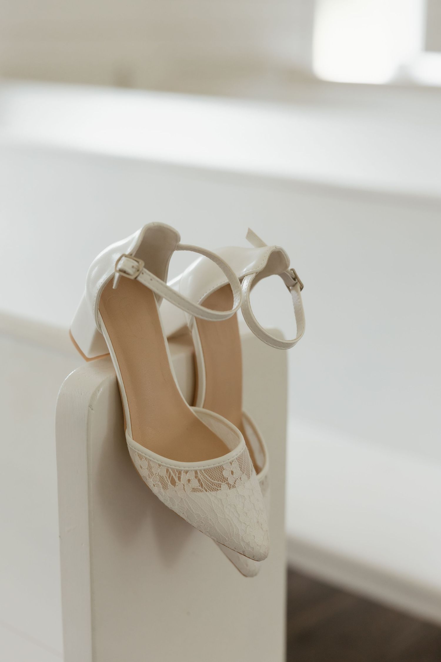 White lace block heel bridal shoes with ankle straps resting on a white church pew.