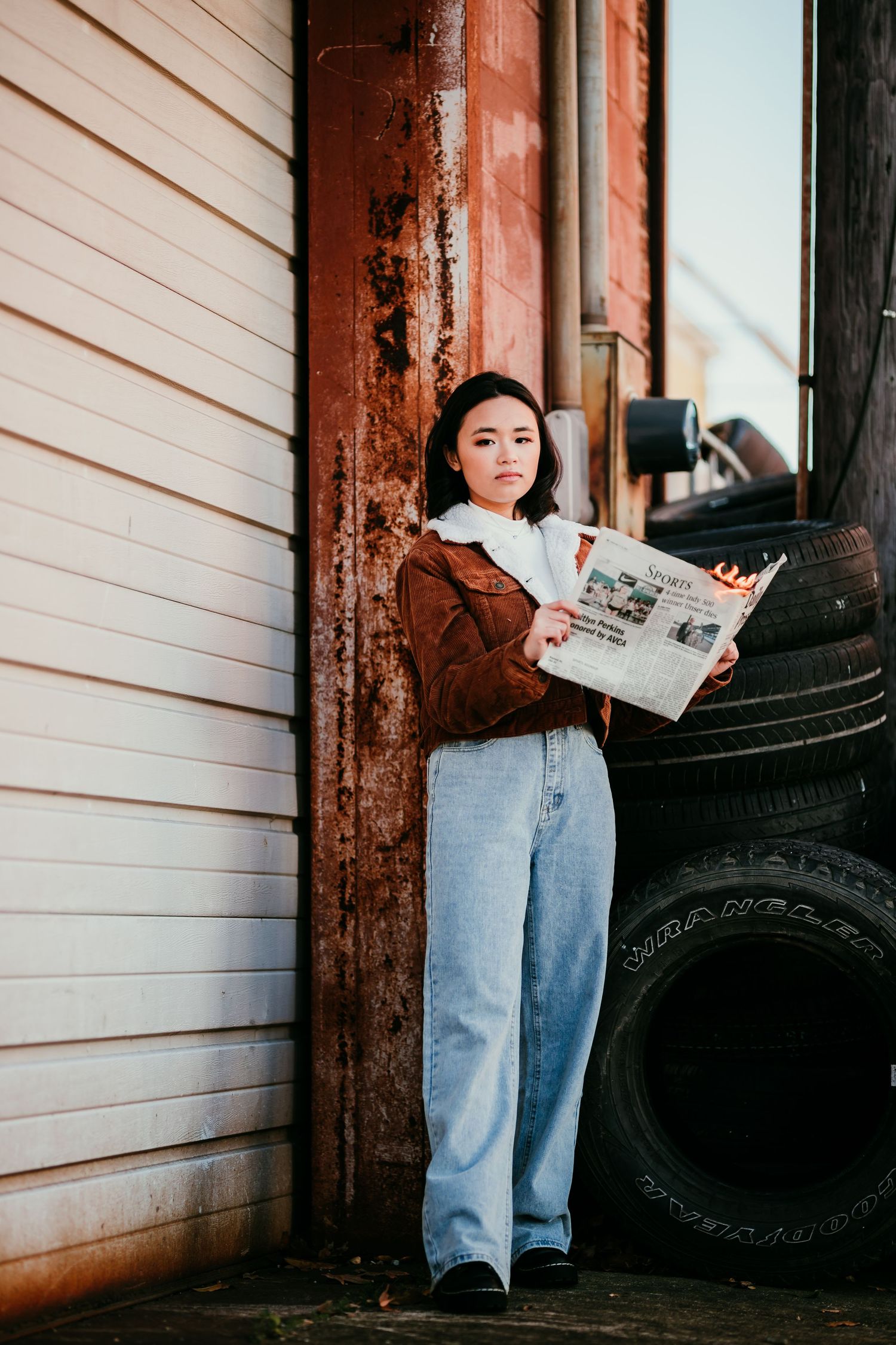 Young Asian woman leaning against rustic building holding newspaper near stacked tires in urban setting.