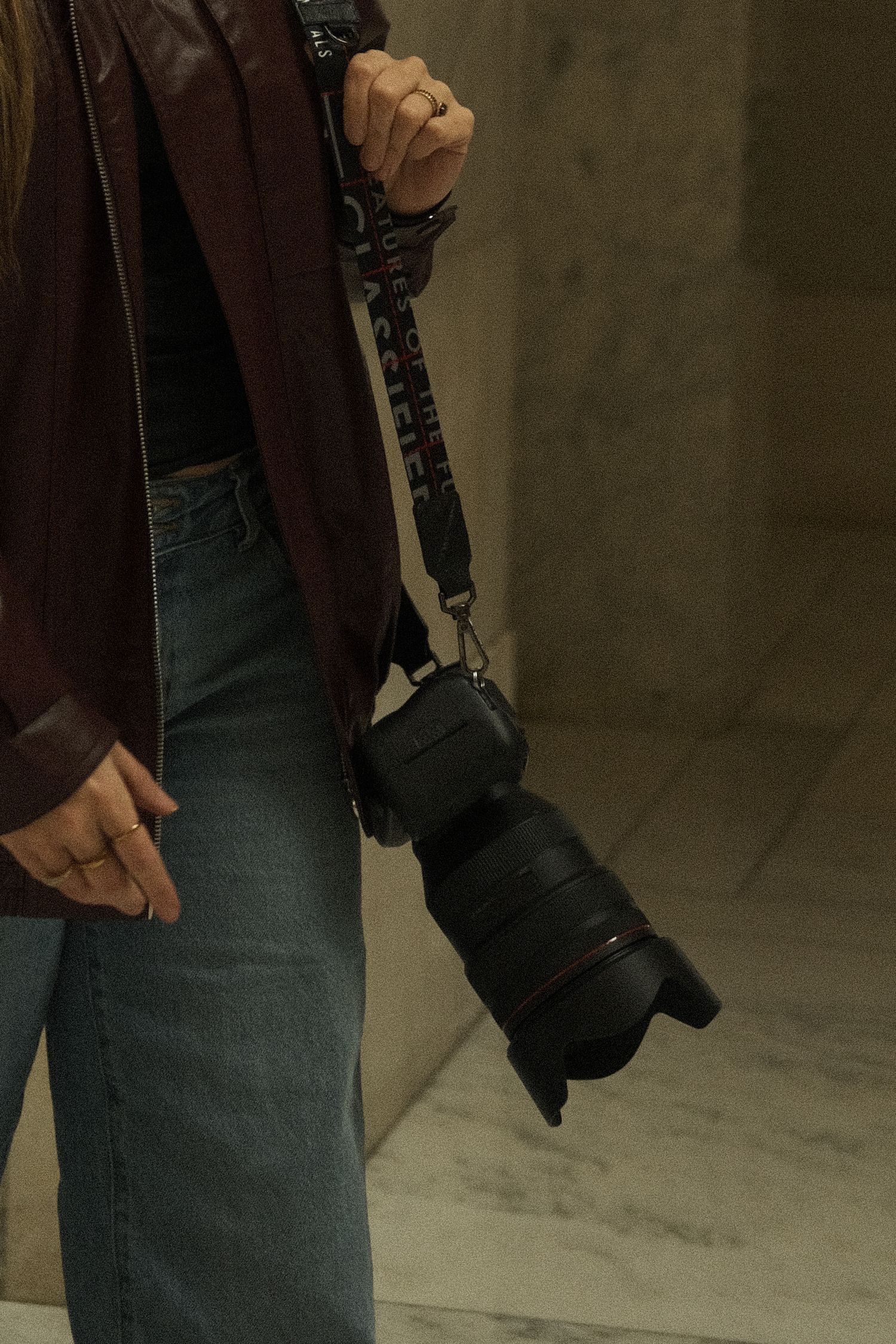 Close-up of woman holding DSLR camera with strap in brown jacket and jeans.