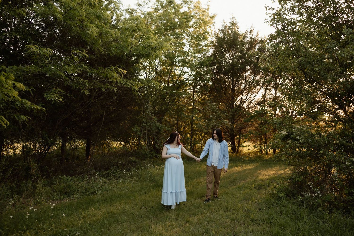 Pregnant couple holding hands walking along a tree-lined path during golden hour sunset.