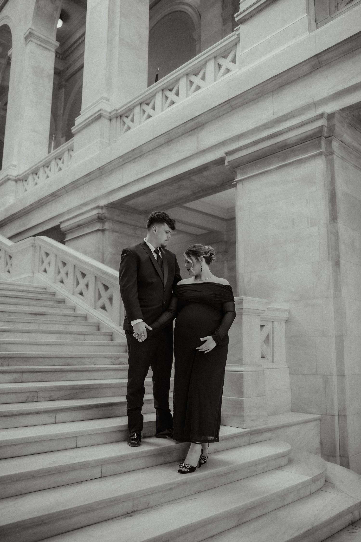 Elegant pregnant couple in formal black attire standing on grand marble staircase, black and white.