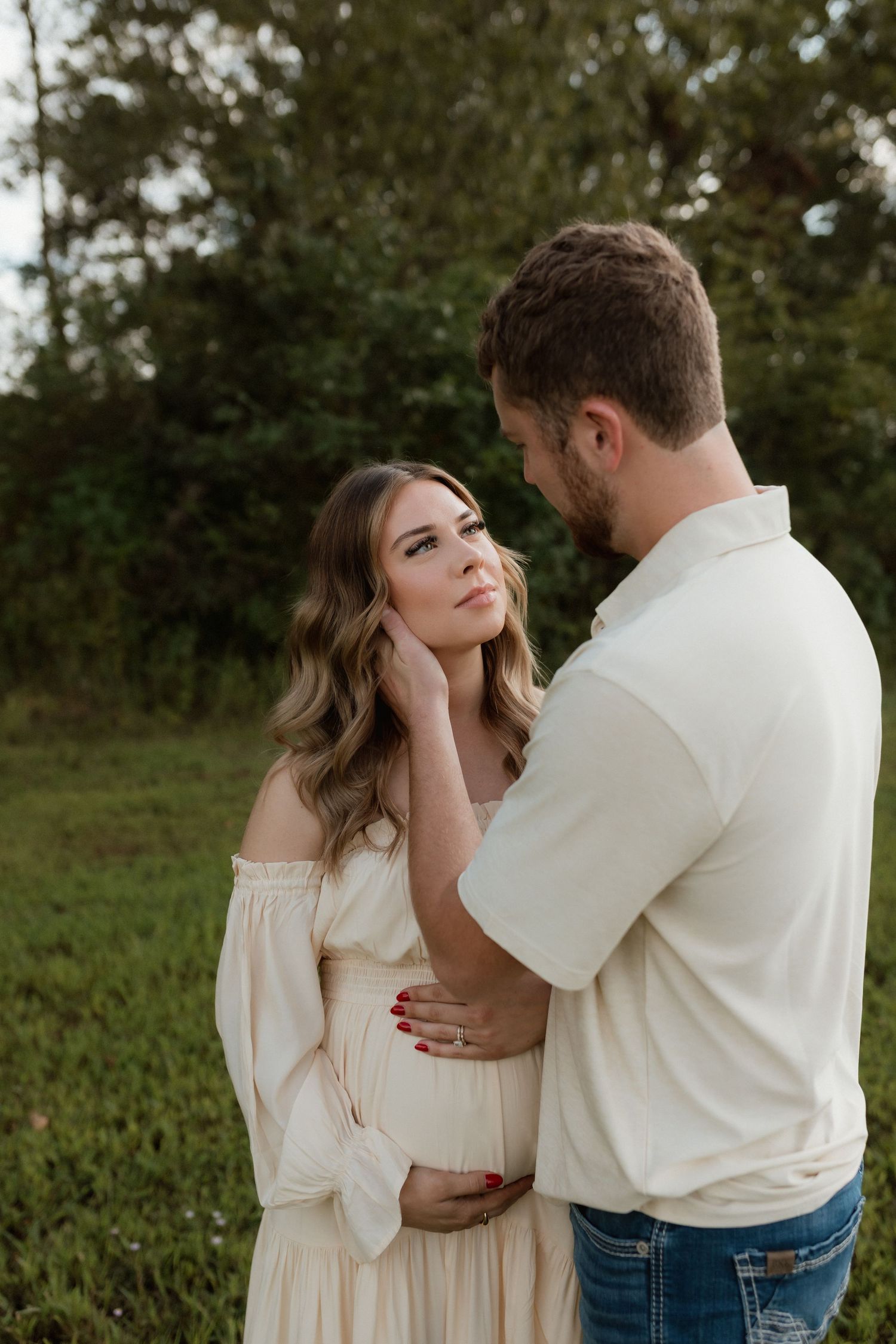 Pregnant woman in cream off-shoulder dress gazing at partner in outdoor maternity photo.