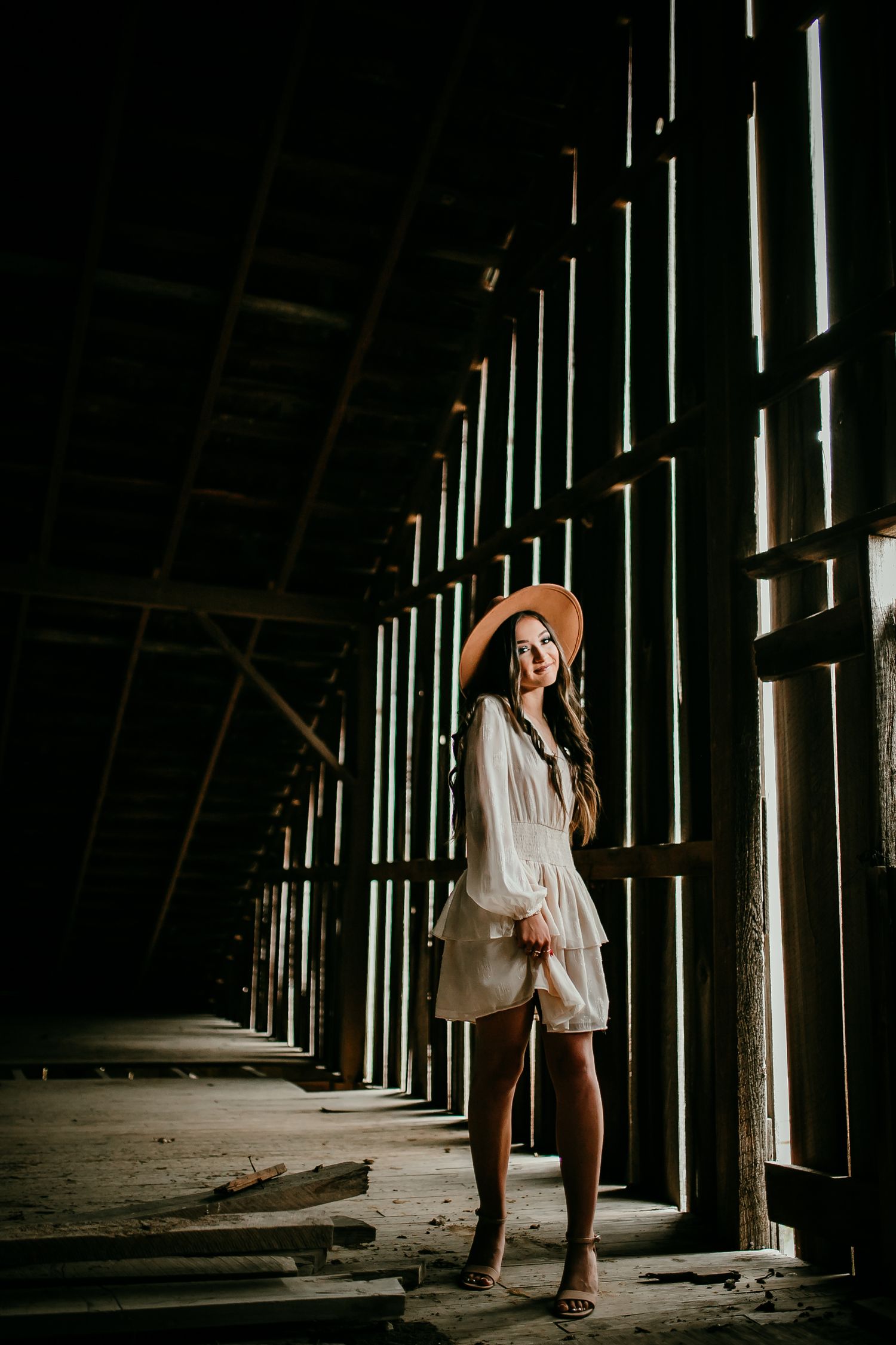 Young woman in white dress and tan hat stands in dramatic light inside rustic wooden barn.