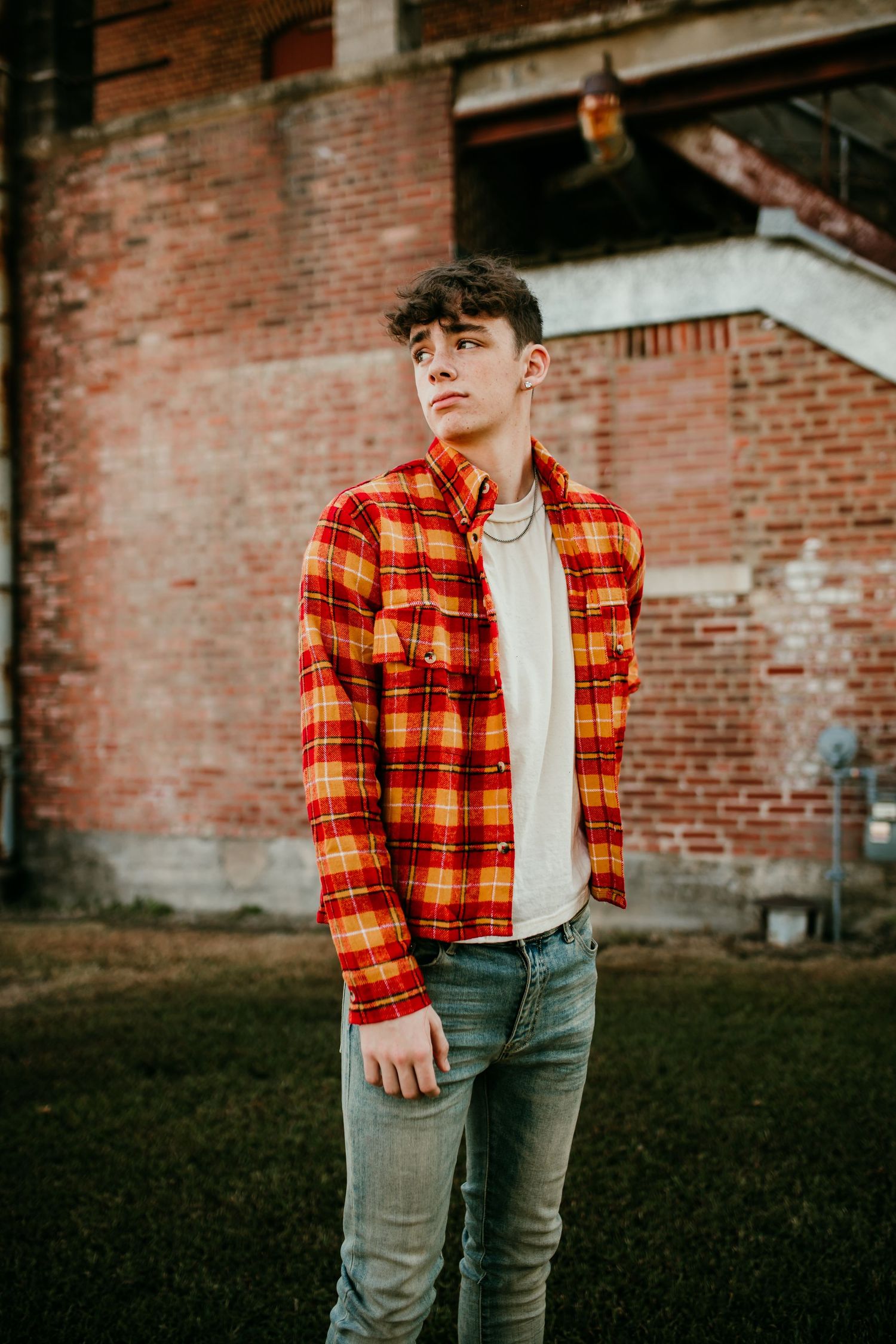 Young man in orange plaid flannel shirt standing before weathered red brick wall.