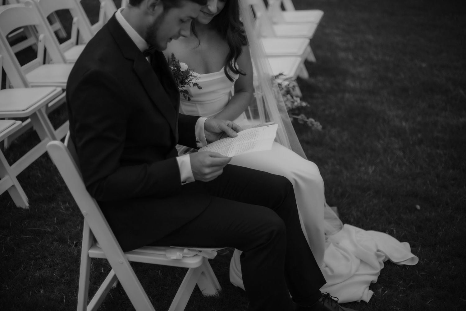 Black and white photo of bride and groom sitting together reading vows at outdoor ceremony.