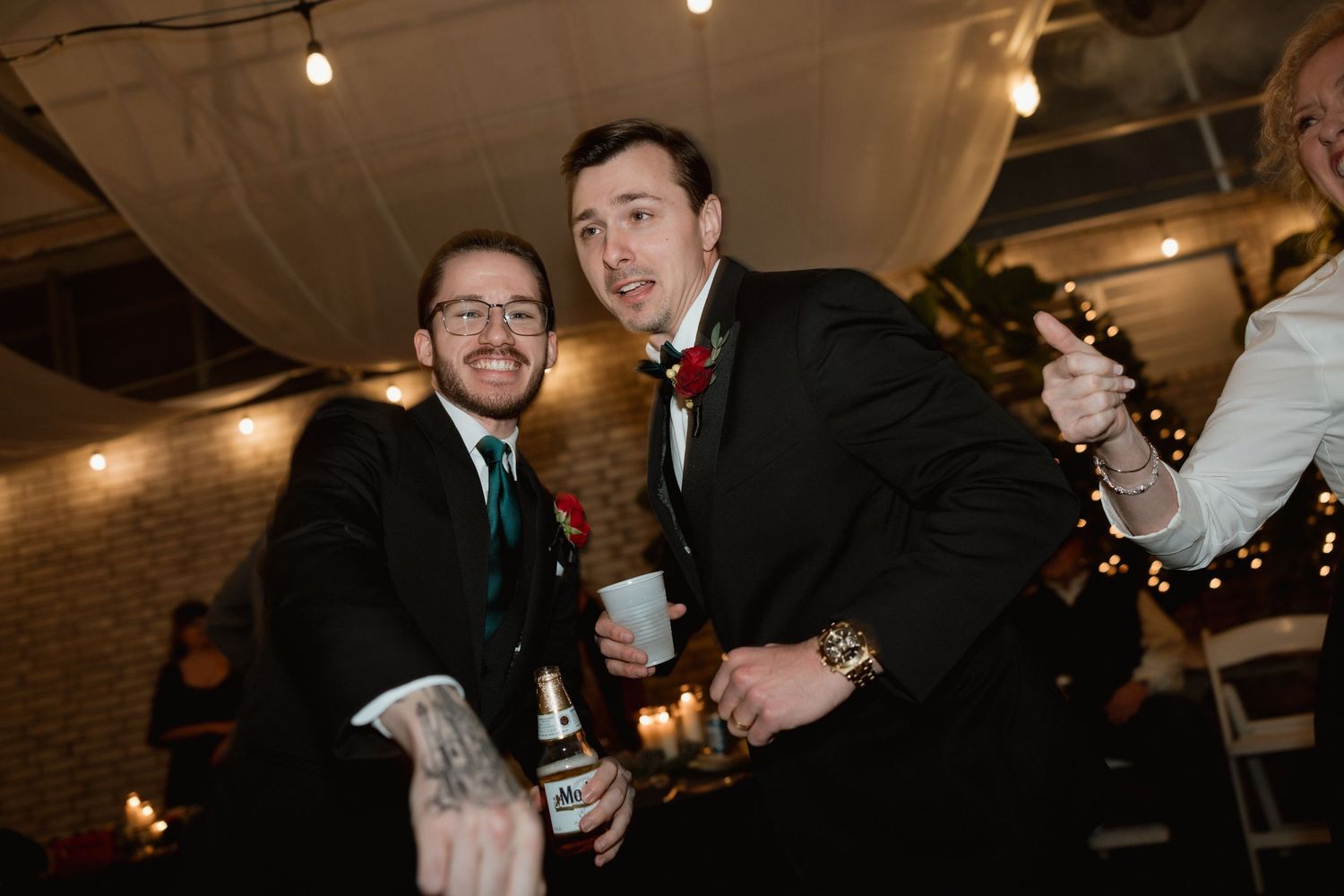 Two groomsmen in black suits celebrating with drinks at wedding reception.