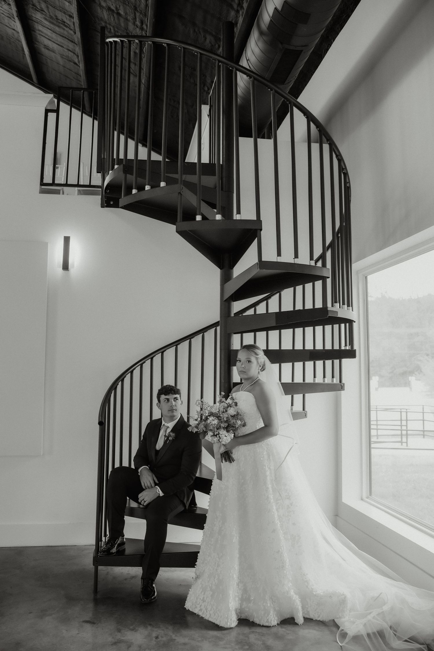 Bride and groom pose elegantly by a black spiral staircase in a black and white wedding portrait.