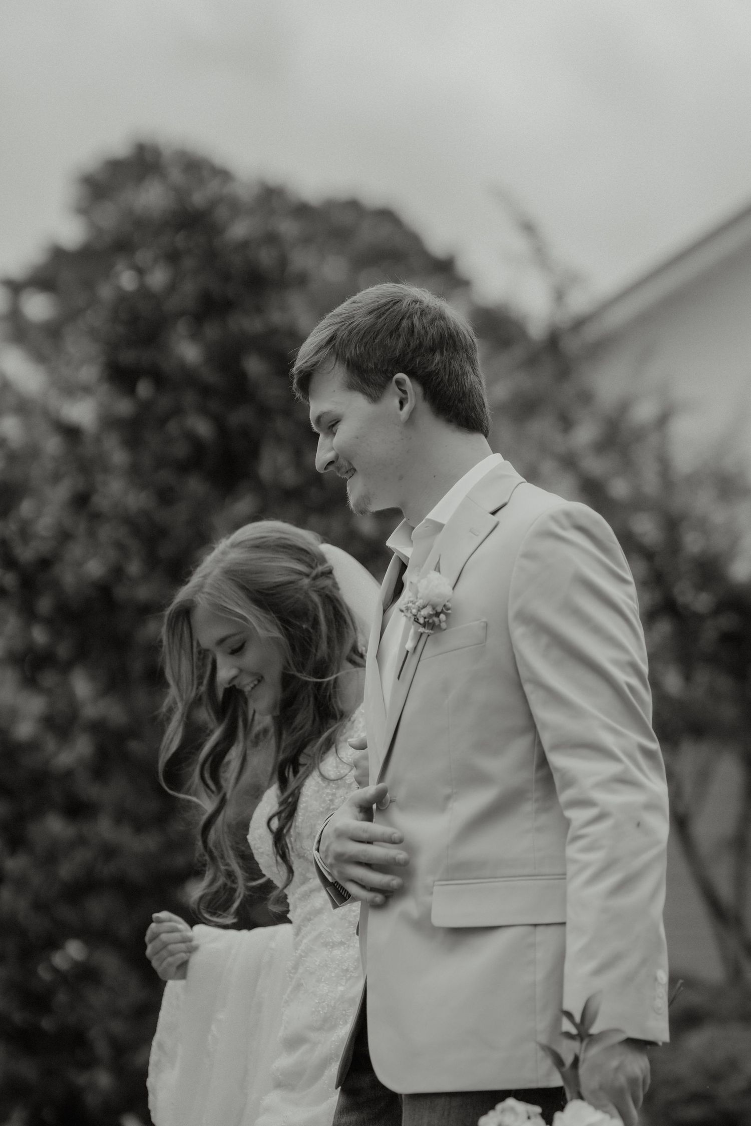 Black and white photo of smiling bride and groom embracing outdoors with lush green trees in the background.