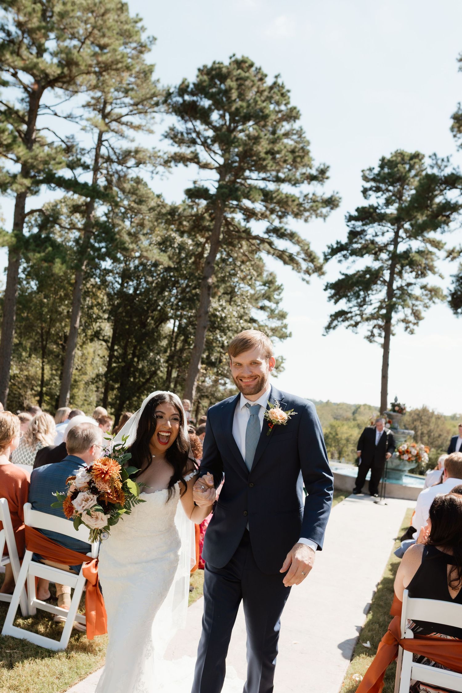 Joyful newlyweds walking down the aisle at an outdoor wedding ceremony surrounded by tall pine trees.