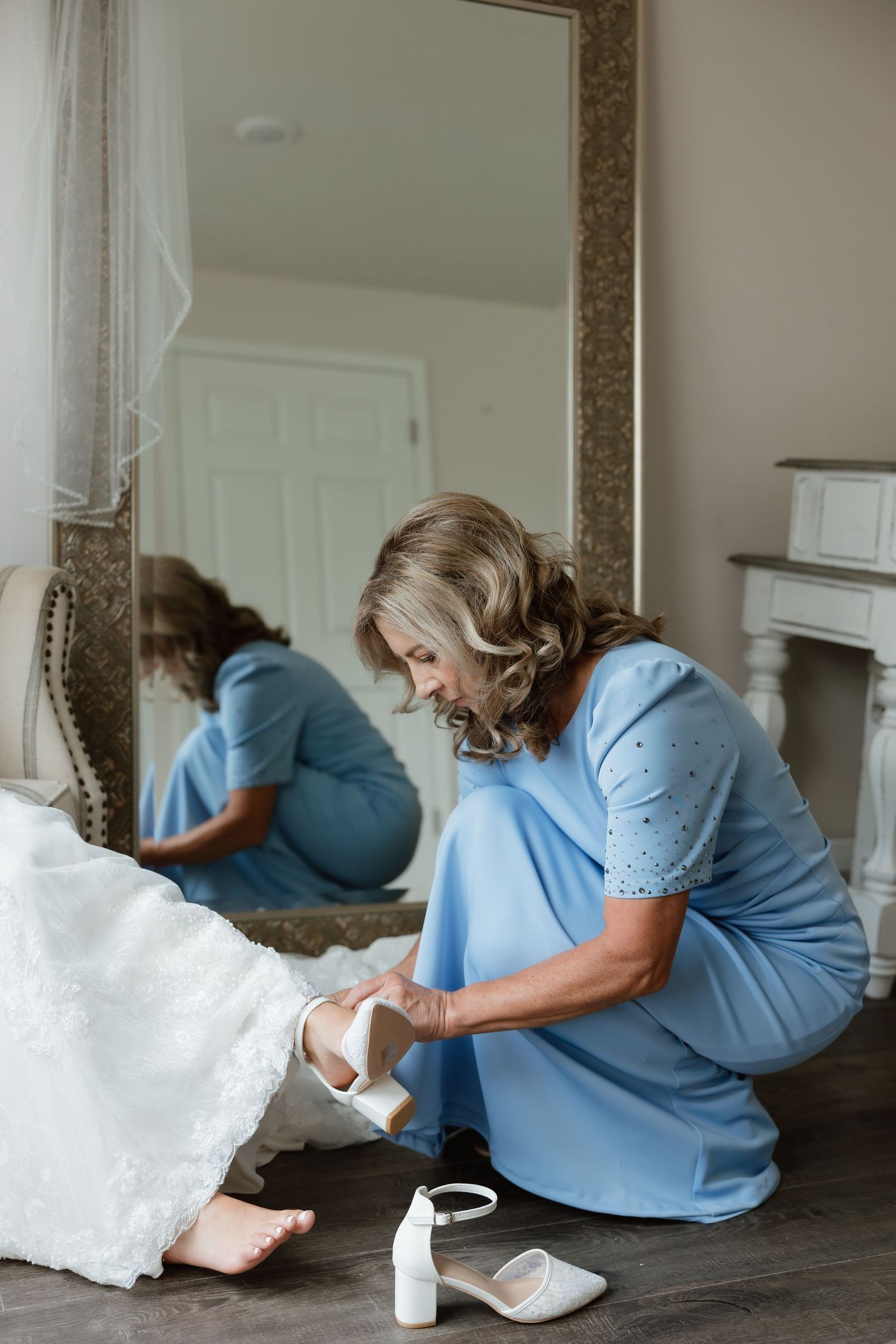 Woman in blue dress helps bride put on white wedding heels in front of a mirror.