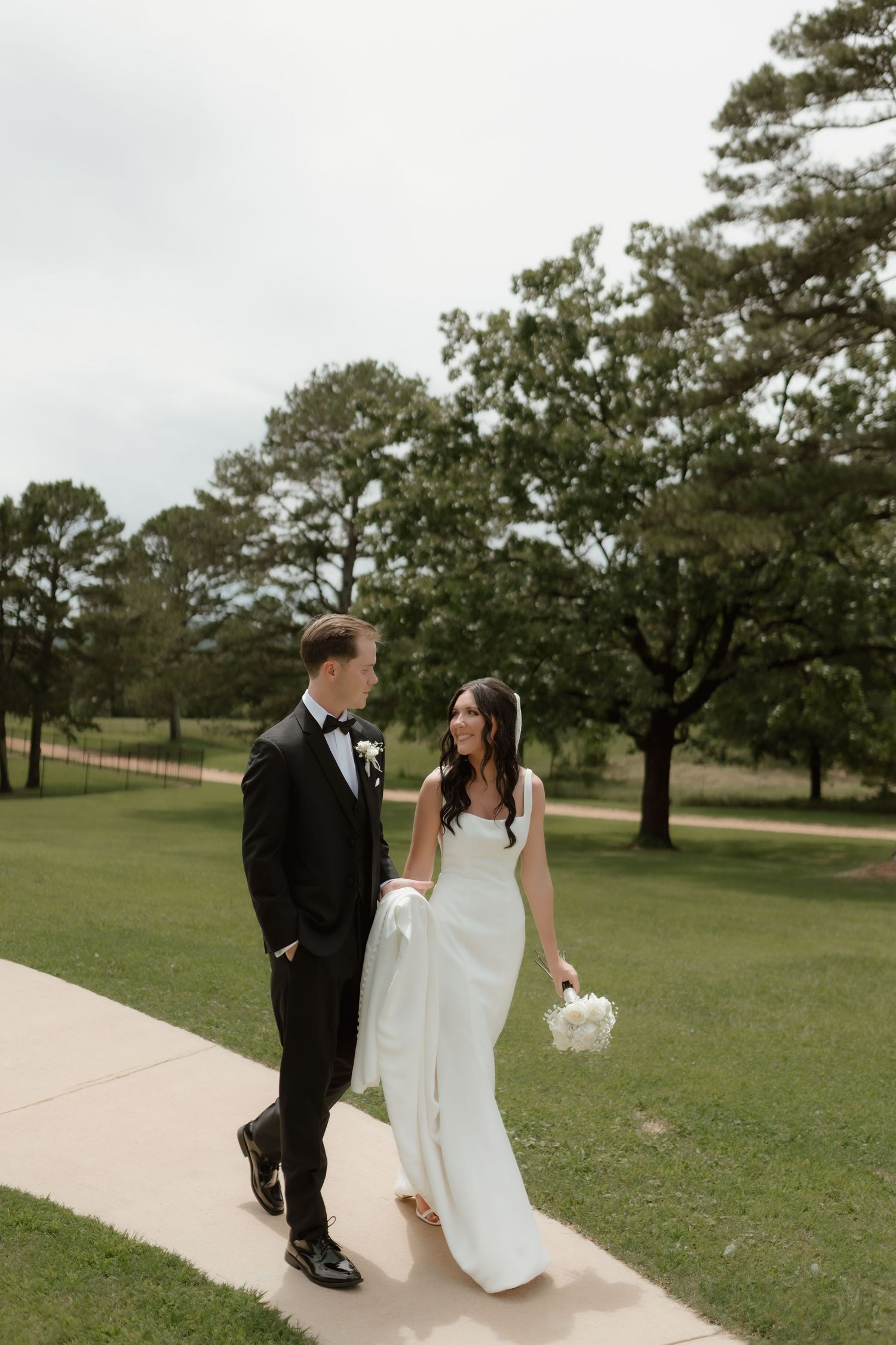 Bride and groom walk hand in hand along a path surrounded by lush green trees on their wedding day.