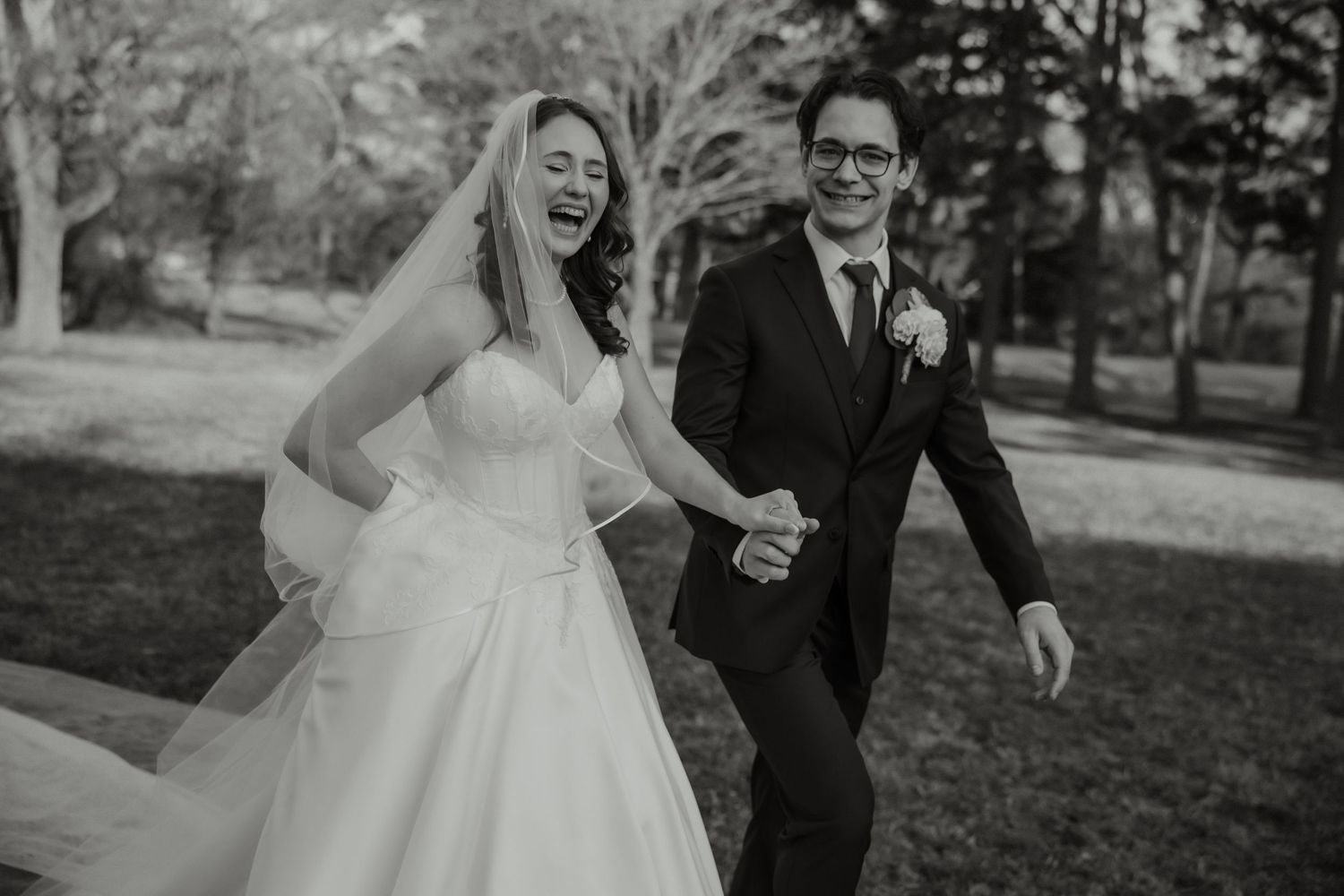 Joyful bride and groom laughing and holding hands while walking outdoors in black and white wedding photo.