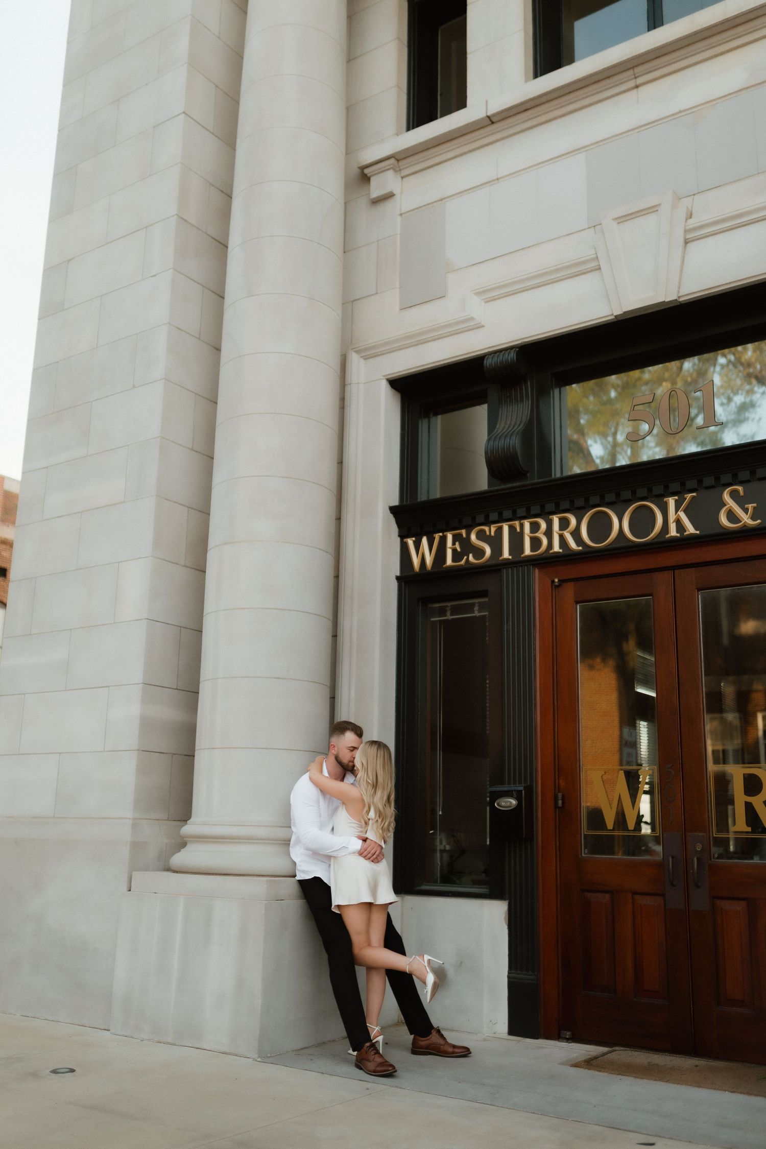 Couple sharing romantic kiss leaning against white stone column outside Westbrook and Reed building entrance.