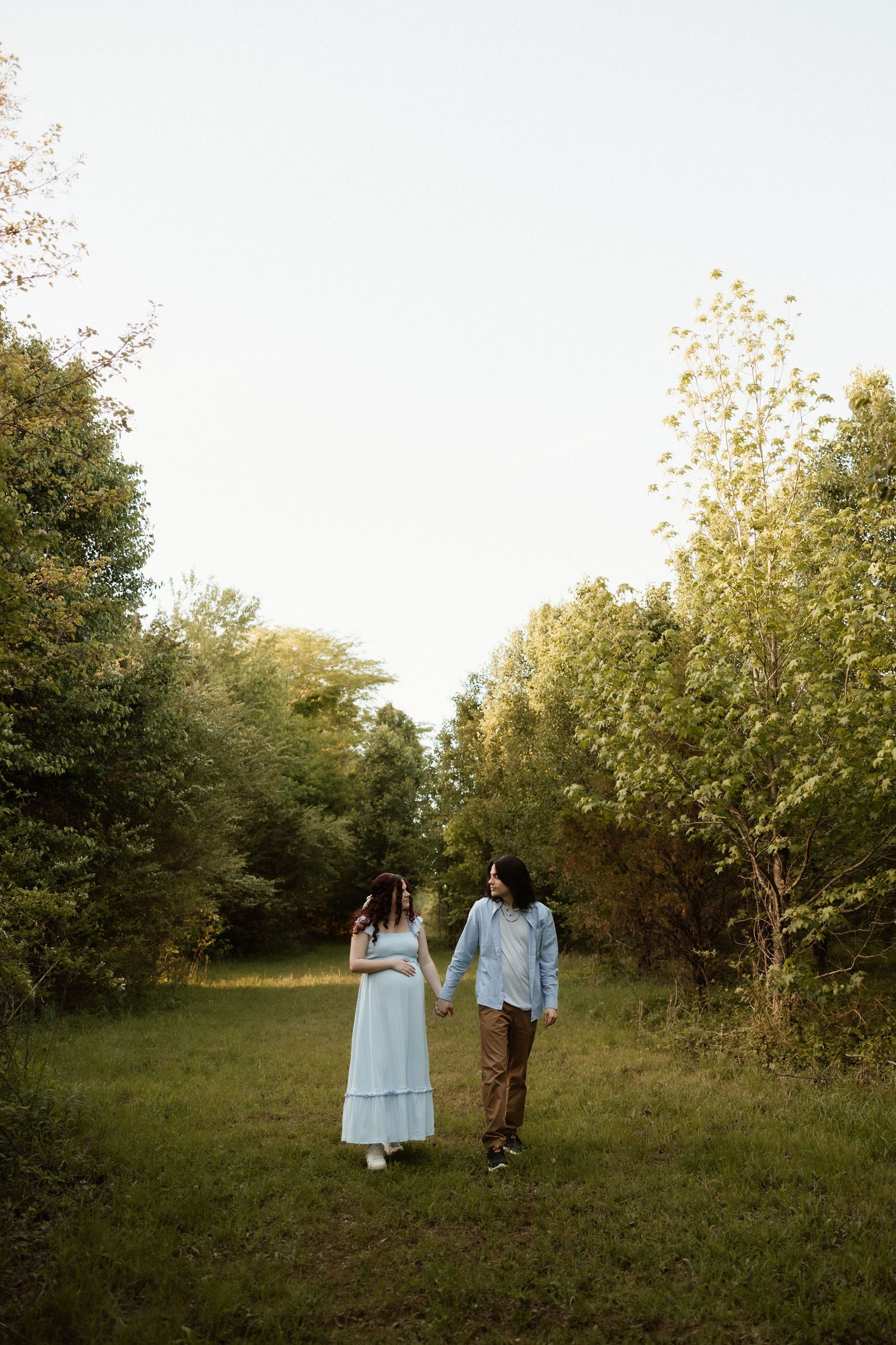 Pregnant couple holding hands walking through a lush green meadow surrounded by trees at golden hour.