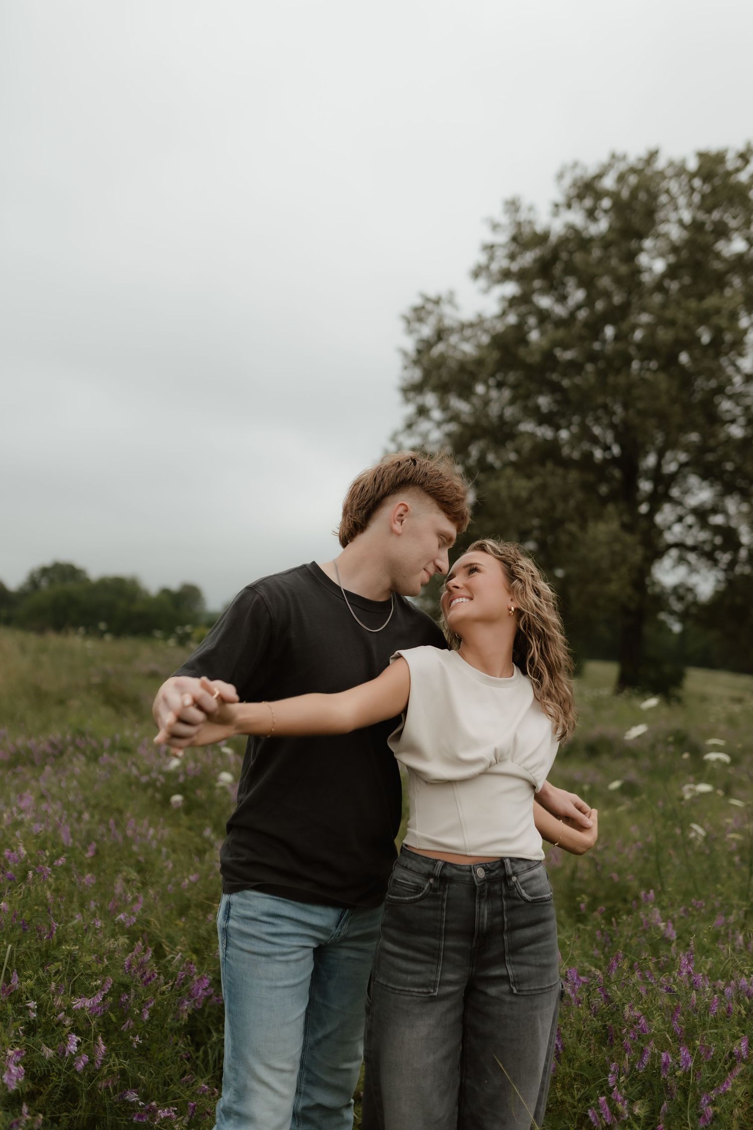 Young couple holding hands and dancing in a purple wildflower field, smiling at each other romantically.