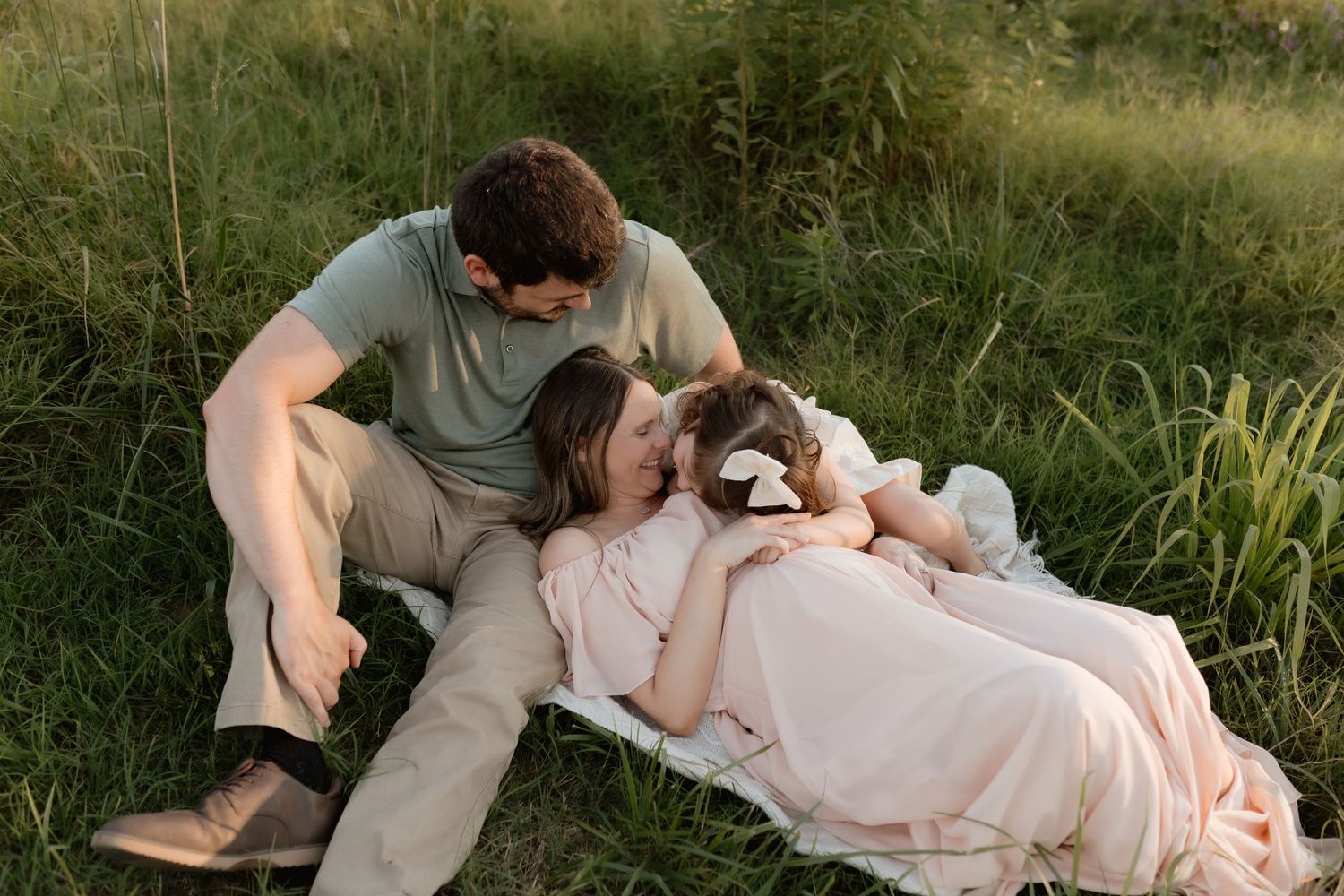 Couple relaxing in green grass with young daughter lying across pregnant mothers lap at golden hour.