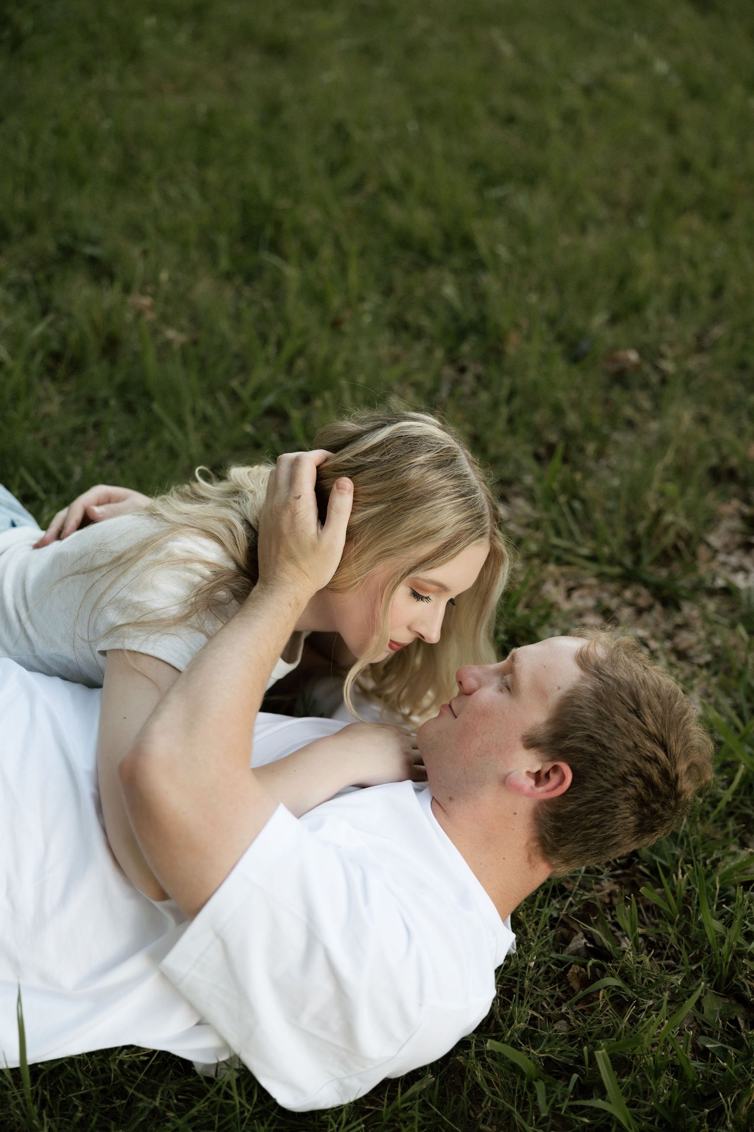 A couple lying on green grass, gazing lovingly at each other during a romantic outdoor engagement session.