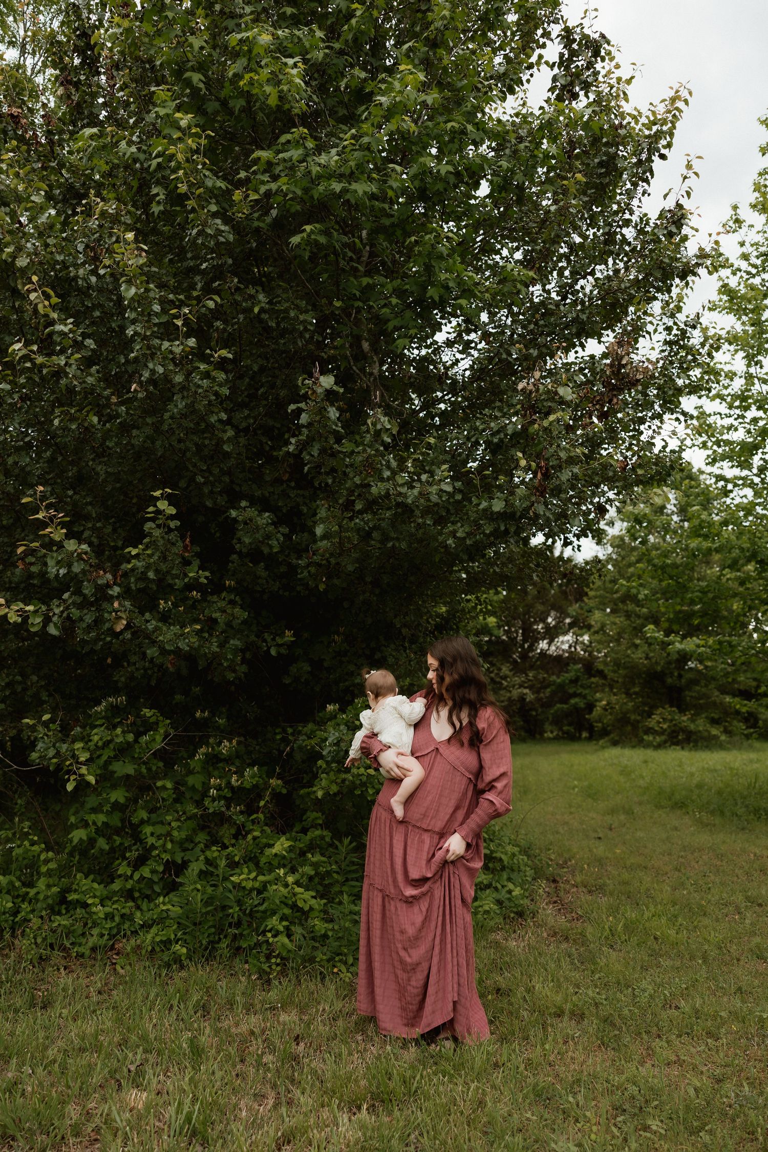 Mother in maxi dress holding baby near large green tree outdoors.