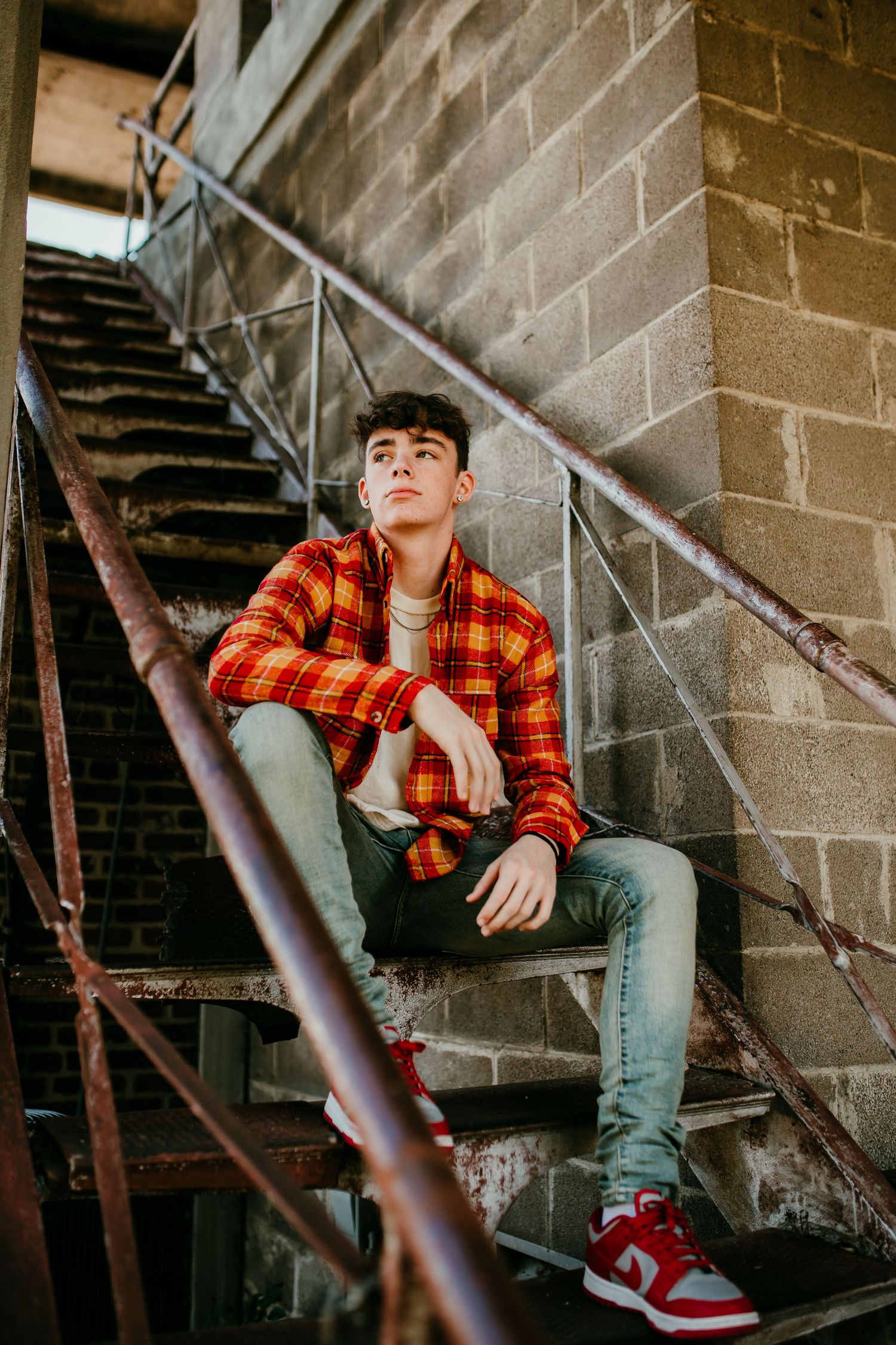 Teen boy in red plaid shirt and jeans sitting on rustic metal fire escape stairs.