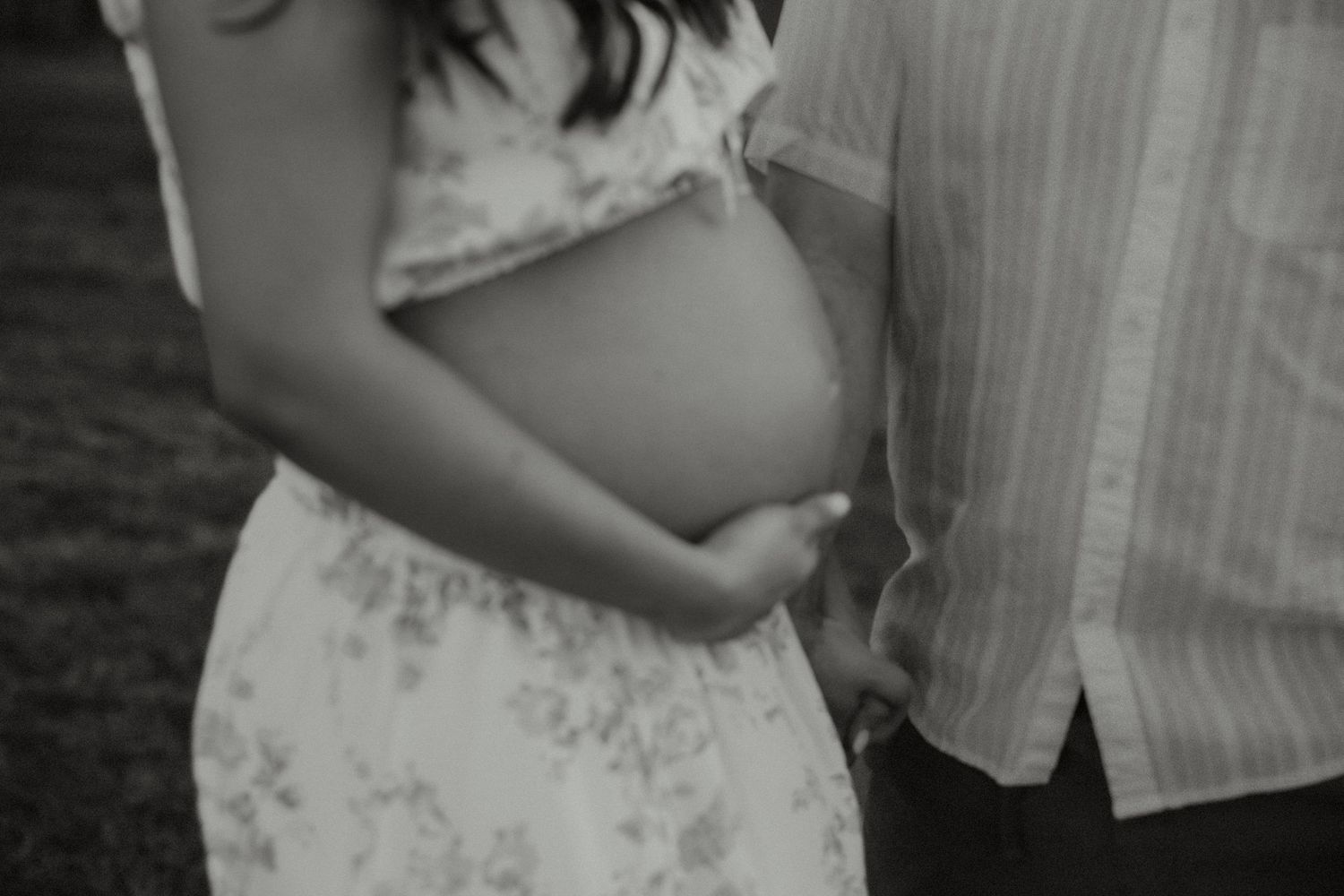 Black and white close-up of a couple cradling a pregnant belly outdoors in a tender maternity portrait.