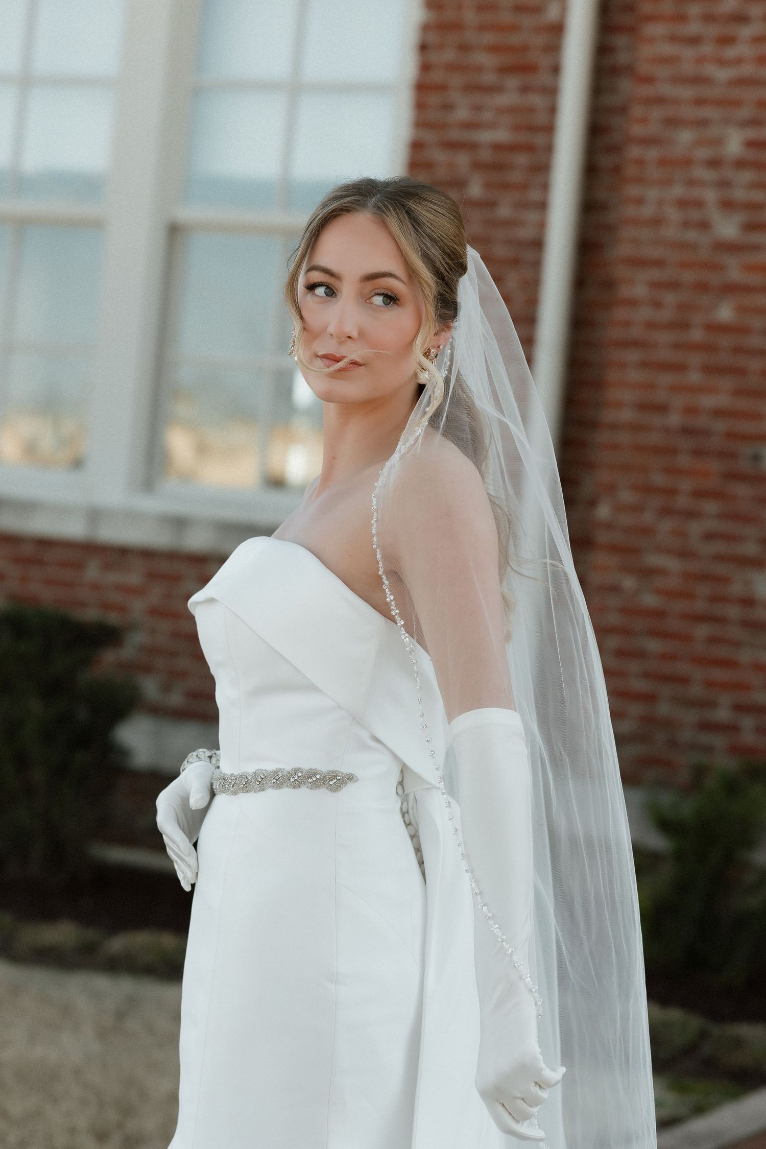 Elegant bride in strapless white gown with long veil and gloves poses outside a brick building.