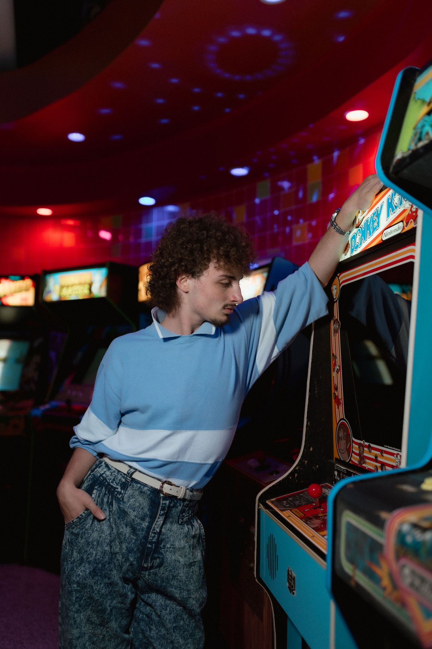 Curly-haired young man in blue polo leans against a vintage arcade game in a neon-lit arcade.