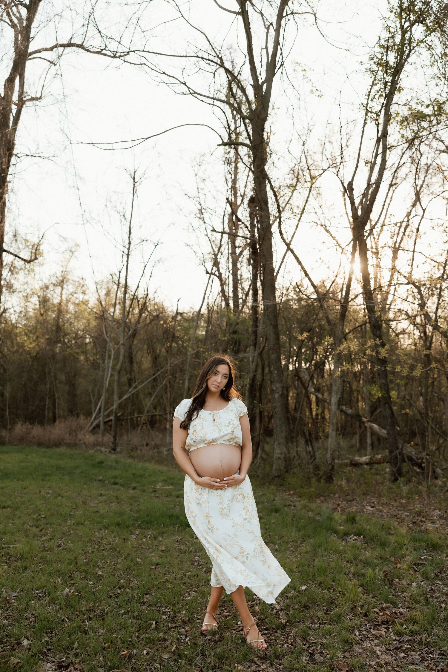 Pregnant woman in a flowing white floral dress cradling her baby bump in a serene wooded outdoor setting at sunset.