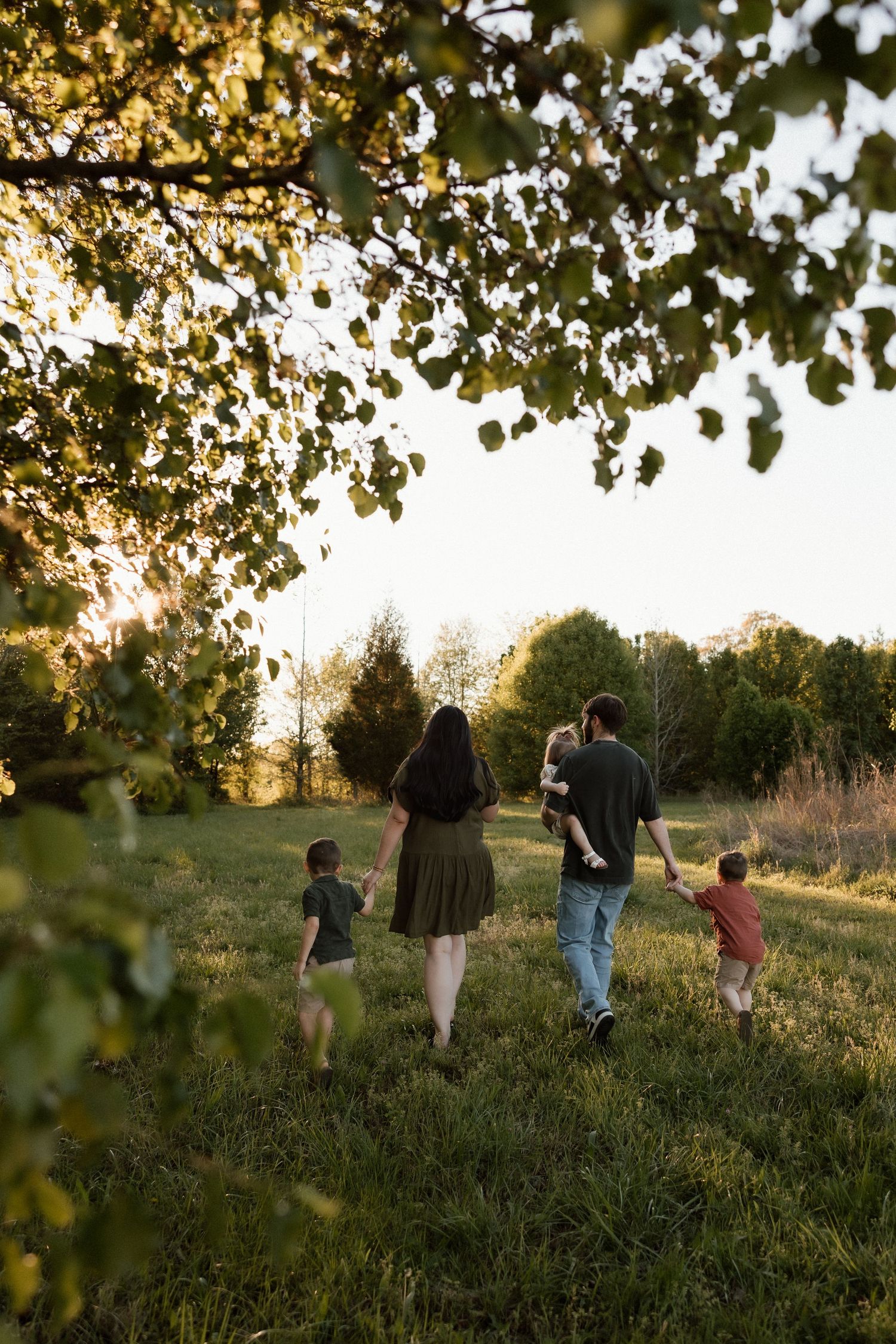 Family of five walking away through a lush green field framed by tree branches at sunset.