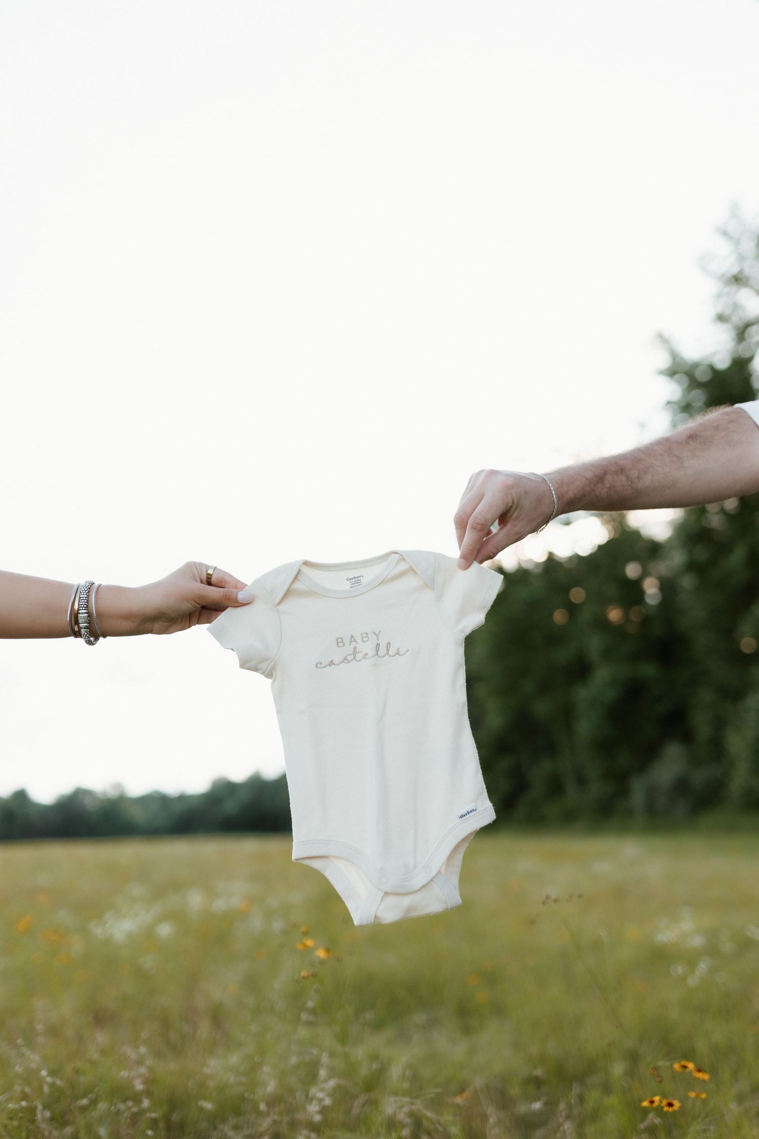 Couple holding a white baby onesie reading Baby Castelli in an open field for pregnancy announcement.