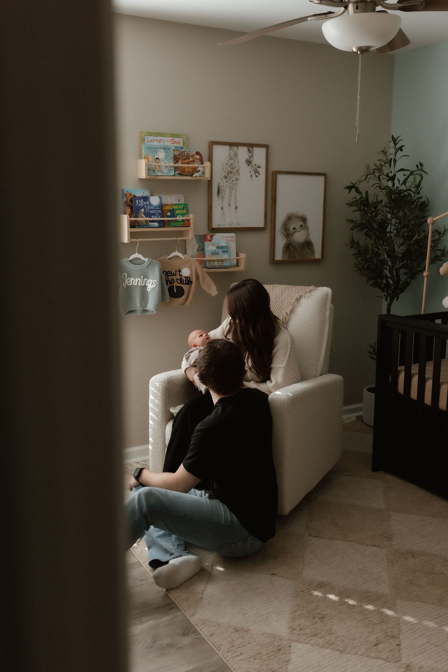 Parents sit together in a beautifully decorated nursery with a glider chair, crib, and gallery wall decor.