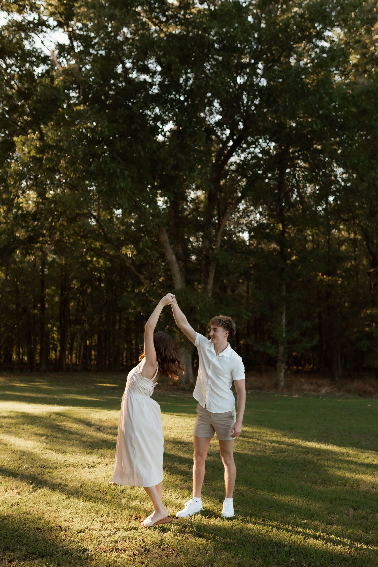 A couple dances together on a green lawn surrounded by tall trees, man spinning woman in white dress at sunset.