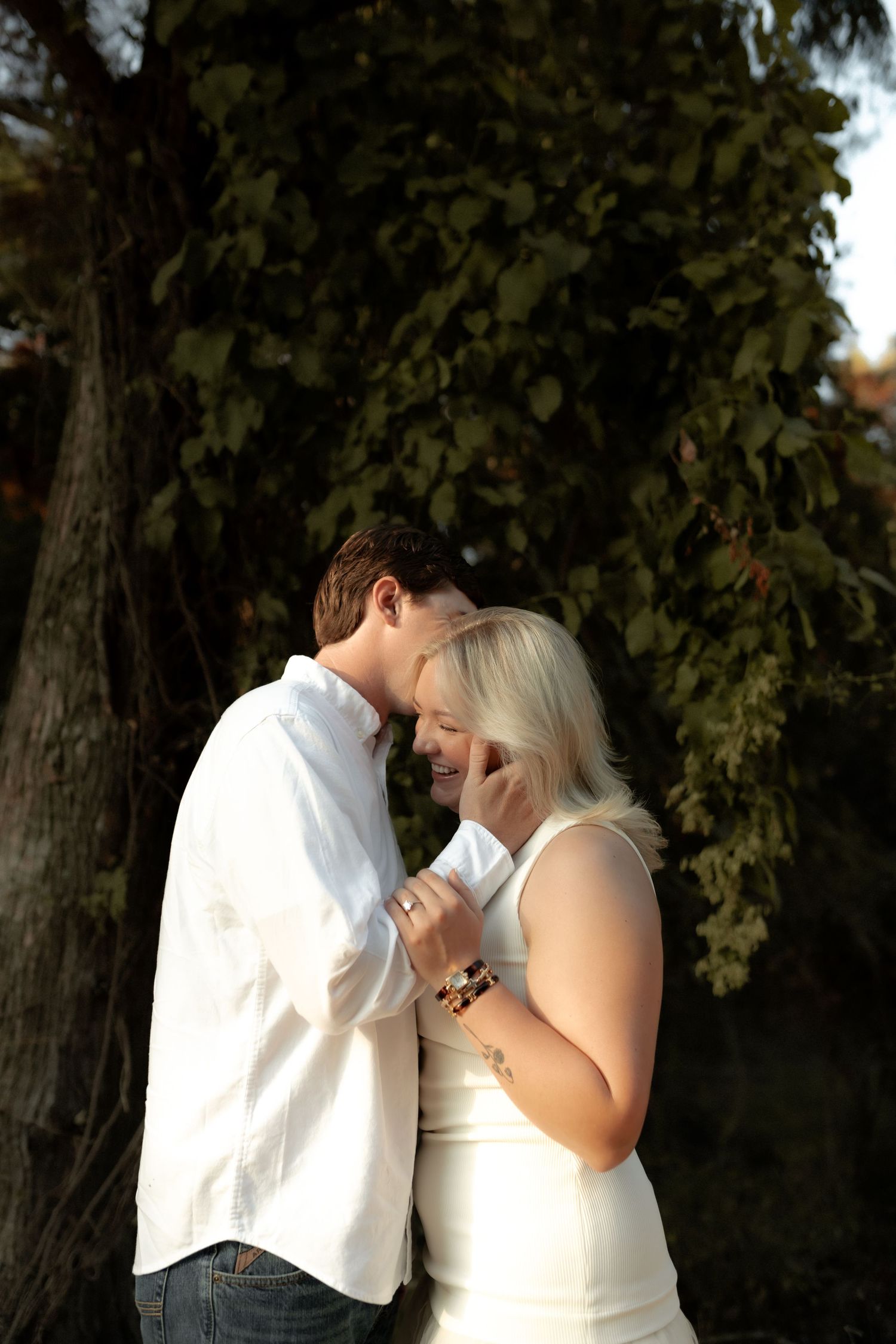Couple in white outfits sharing an intimate moment under a large tree in warm evening light.