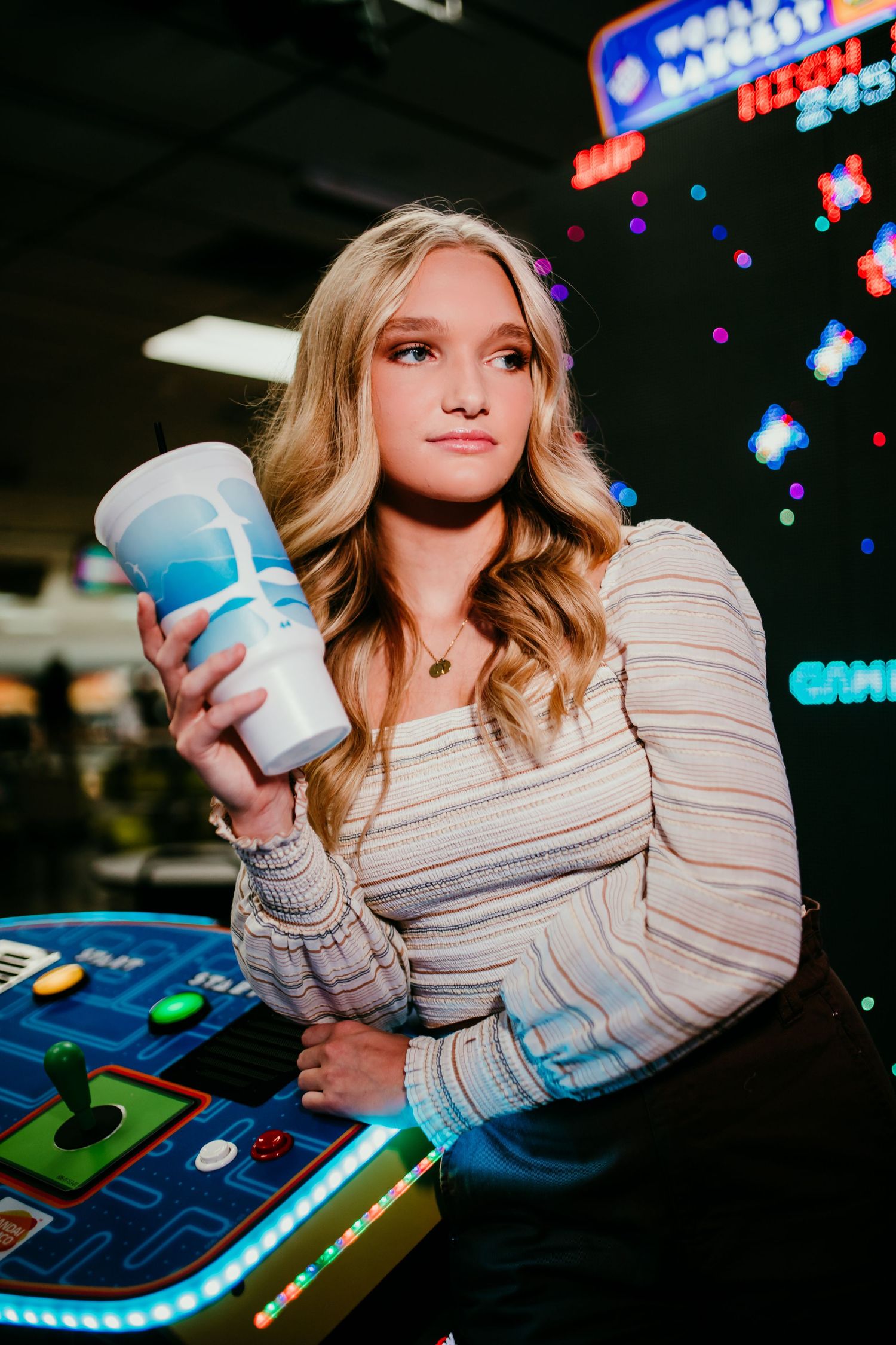 Blonde teen holding a drink cup at a colorful arcade.